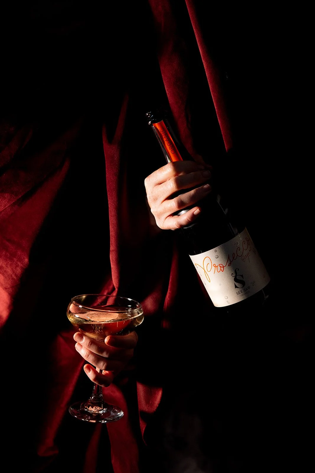 Person in a red velvet blazer holding a glass of champagne and a bottle of Prosecco in a dark setting with minimal lighting.