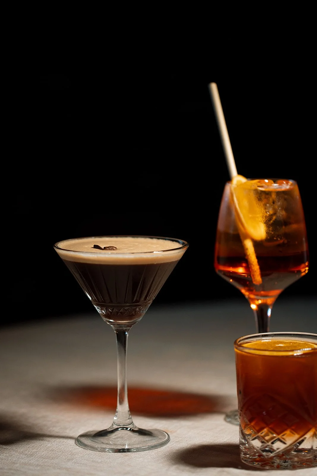Three cocktails on a table against a black background. The front drink is a with a creamy beige froth and dark chocolate shavings. The middle drink is red with lemon slices and a straw. The right drink is amber-colored in a short glass.