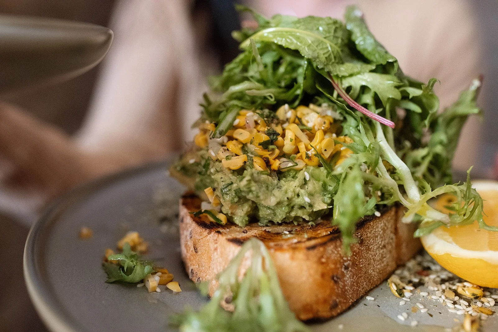 Avocado toast topped with mixed greens, chopped corn, and a lemon wedge, served on a gray plate.