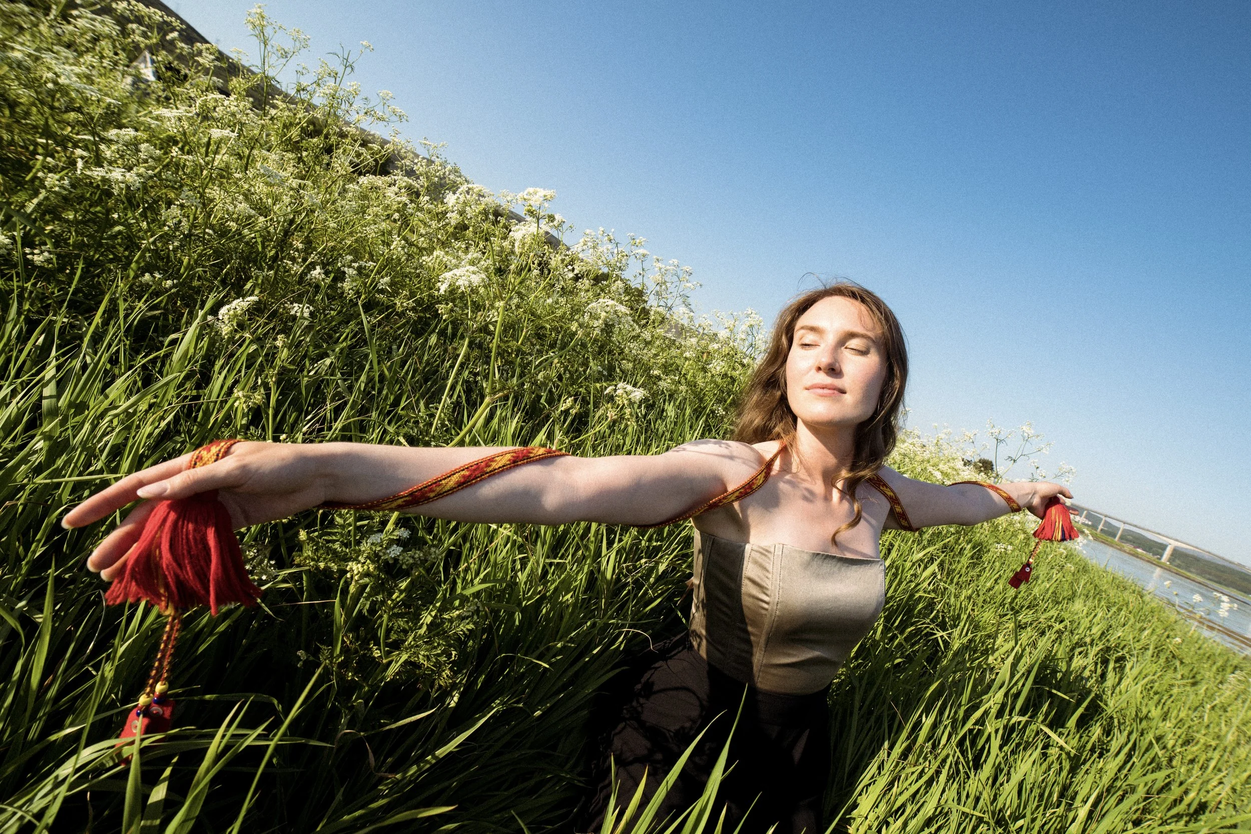 A woman with long brown hair stands with arms outstretched in a grassy field with white wildflowers, eyes closed and face relaxed, under a clear blue sky, near a body of water, with a bridge in the distance.