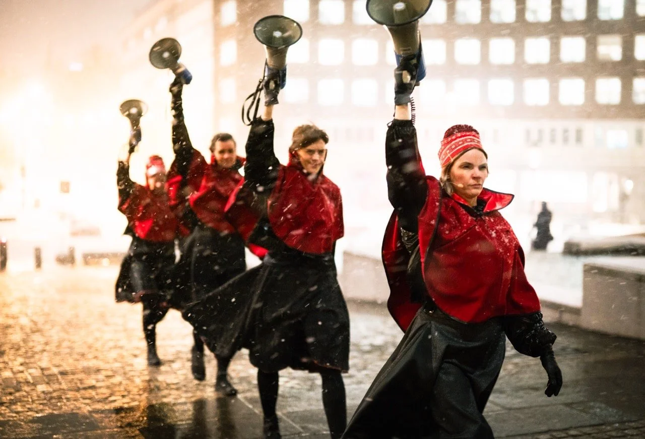 Four women dressed in red and black outfits holding megaphones on a city street at night, possibly protesting or participating in a demonstration.
