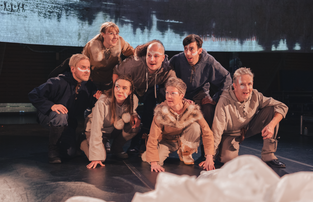 A group of seven actors in costume posing on a stage, with a backdrop depicting a lake and trees.
