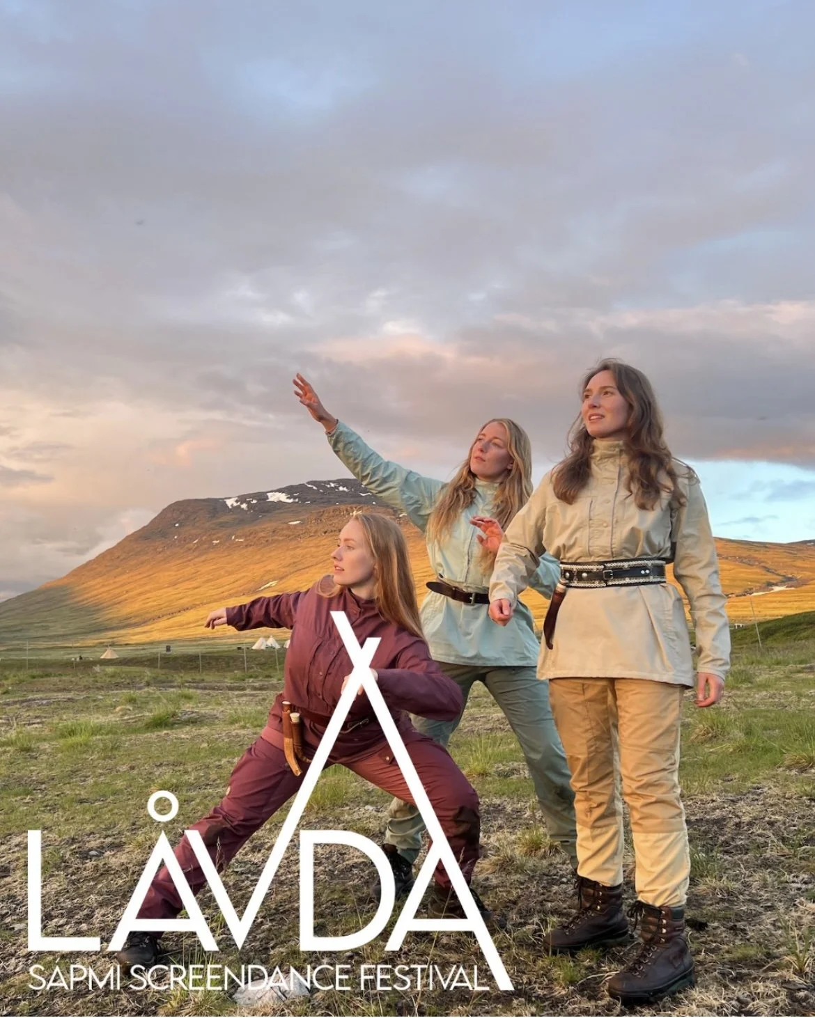 Three women dressed in outdoor clothing standing on a grassy field with mountains in the background, one is kneeling and pointing, another is posing with a hand raised, and the third is standing smiling. The image promotes the Lávída Sapmi ScreenDance Festival.