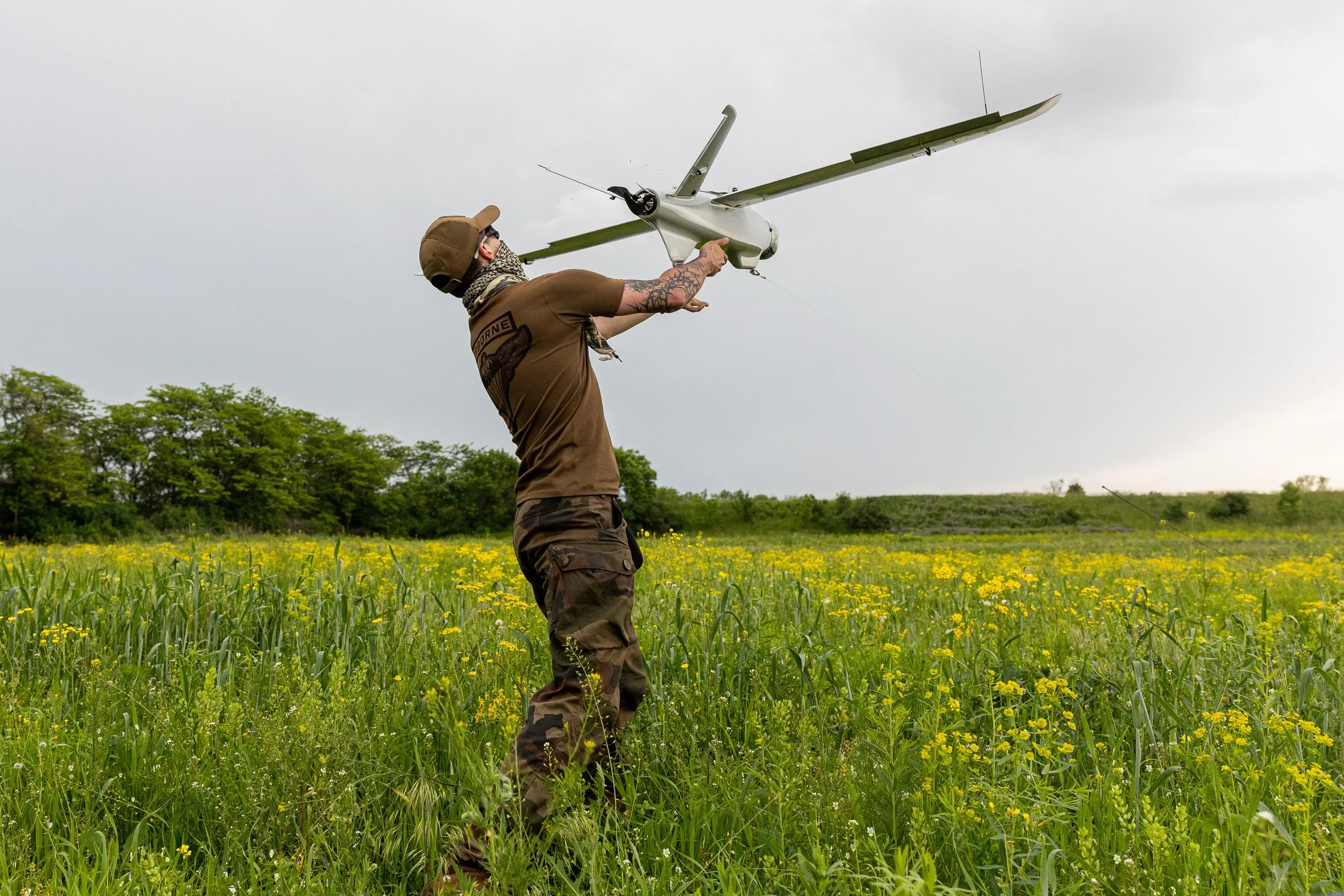 Person in military clothing launching a small drone in a grassy field with yellow flowers.