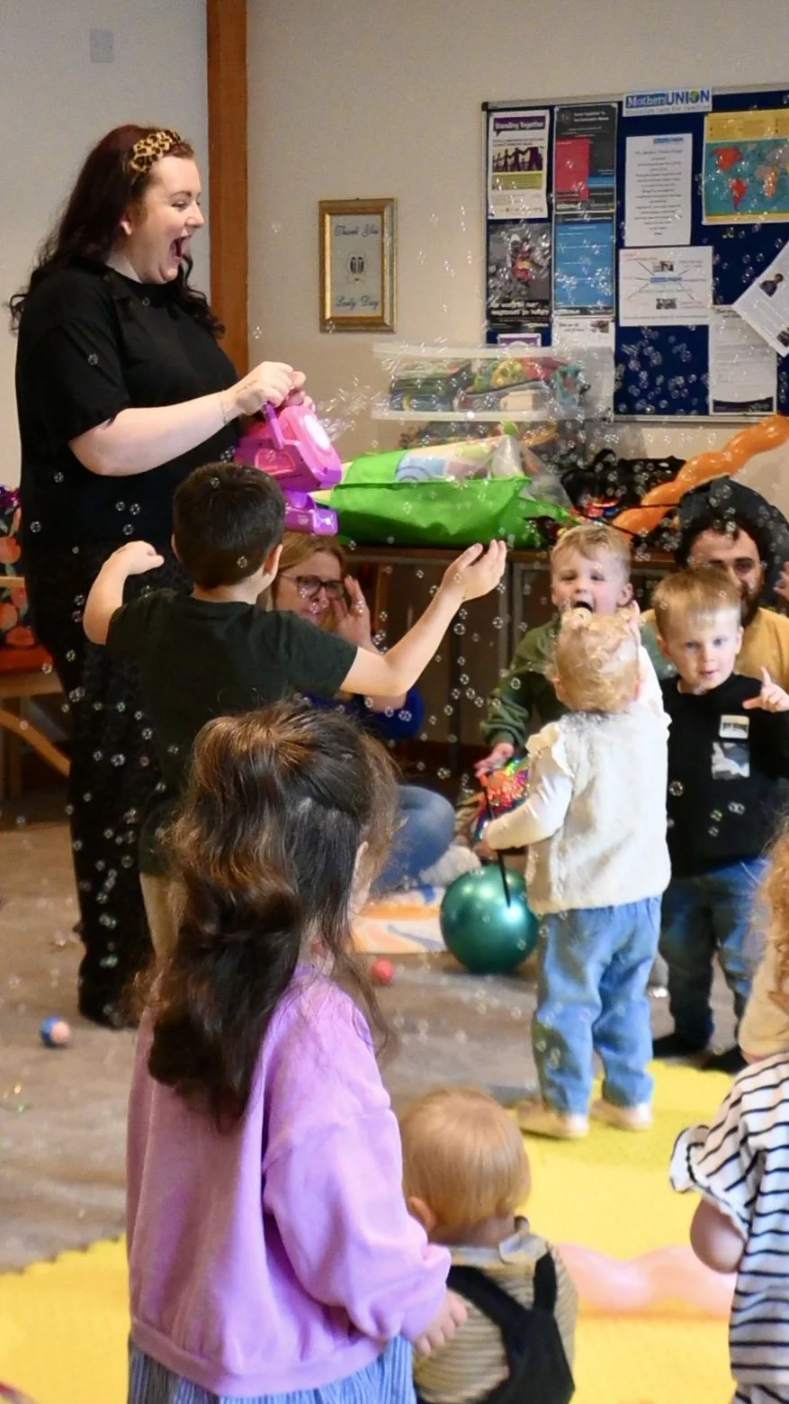 teacher vikki with a bubble machine blowing bubbles to class of smiling toddlers and pre-schoolers