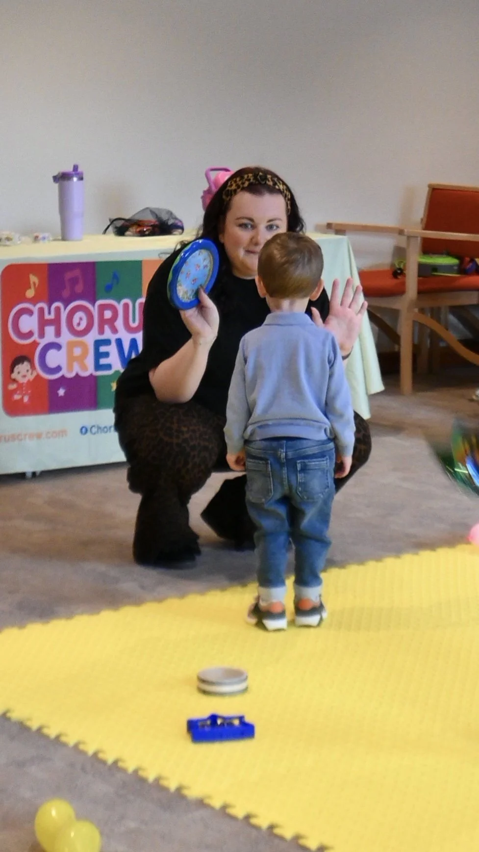 Teacher Vikki with a small boy dressed in blue playing with a tambourine in music class