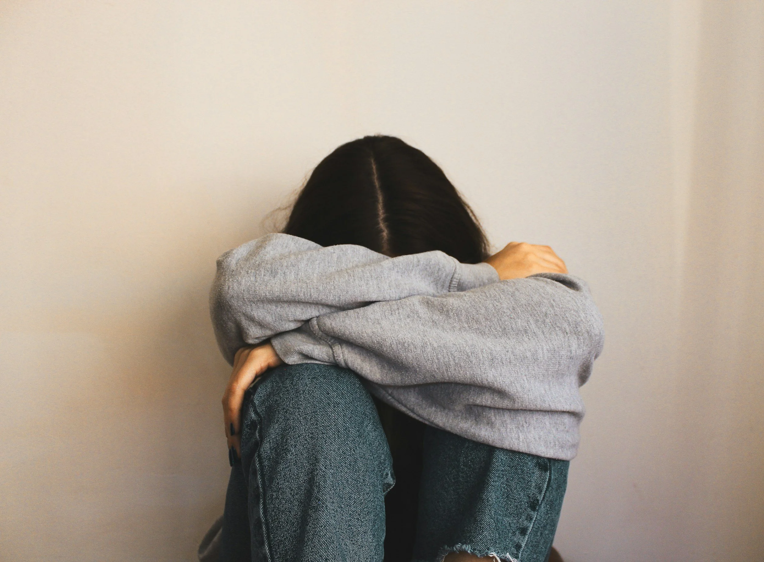 A person with long dark hair, wearing a gray sweatshirt and jeans, sitting on the floor against a plain white wall with their knees drawn up and head resting on their arms, appearing to be upset or distressed.