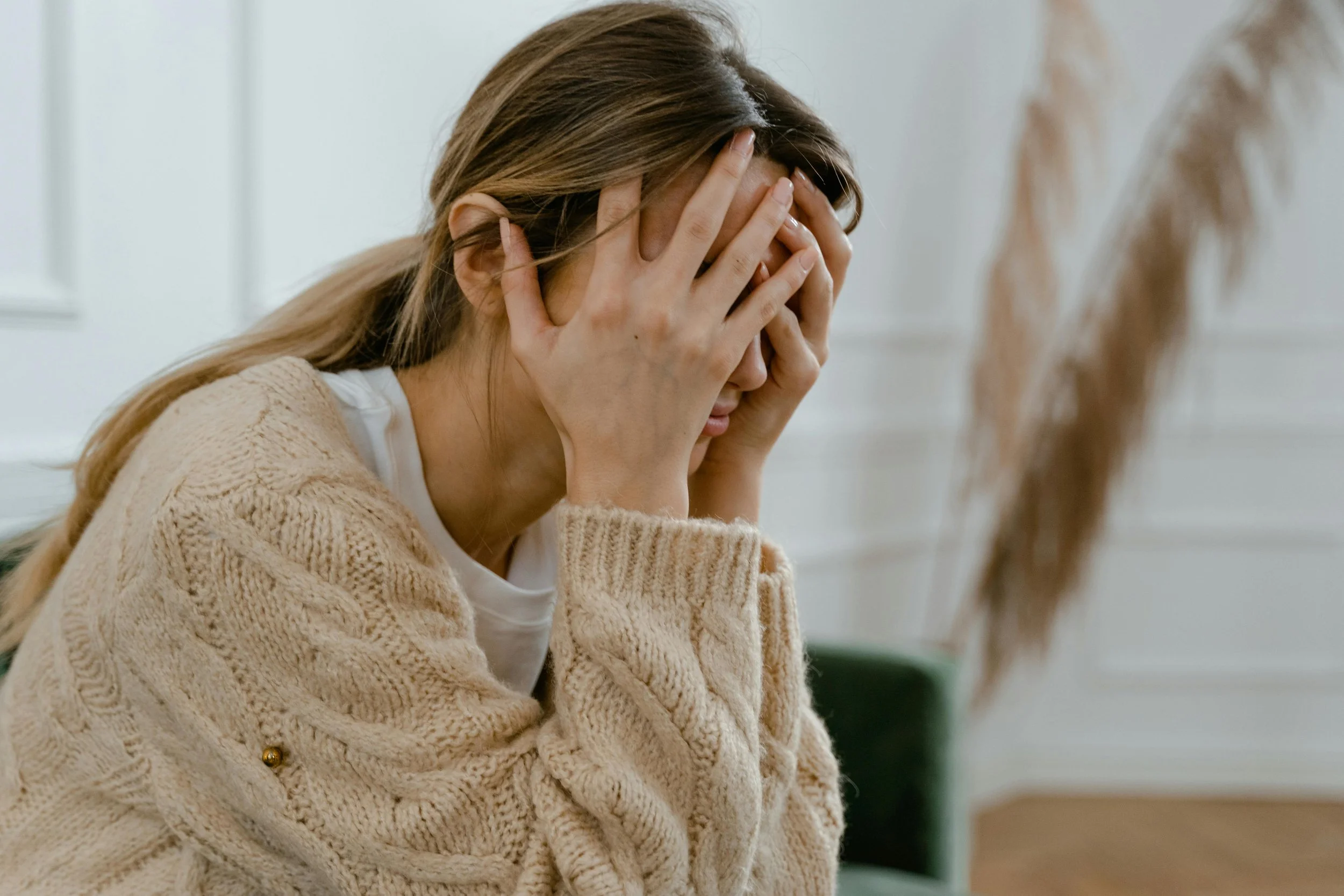 A woman with long hair, wearing a beige knitted sweater, sits with her hands covering her face in distress in a room with white walls.