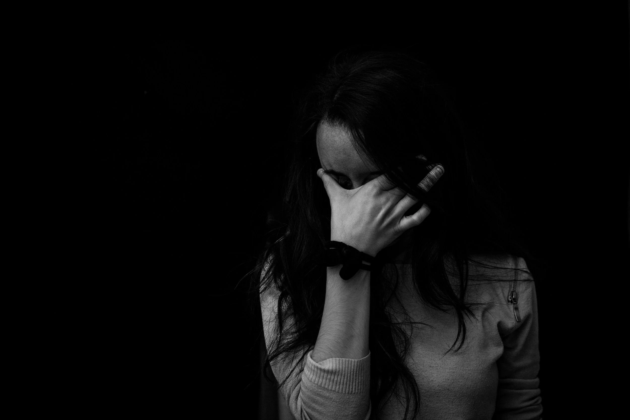 A woman with long hair covering her face with her hand against a black background.