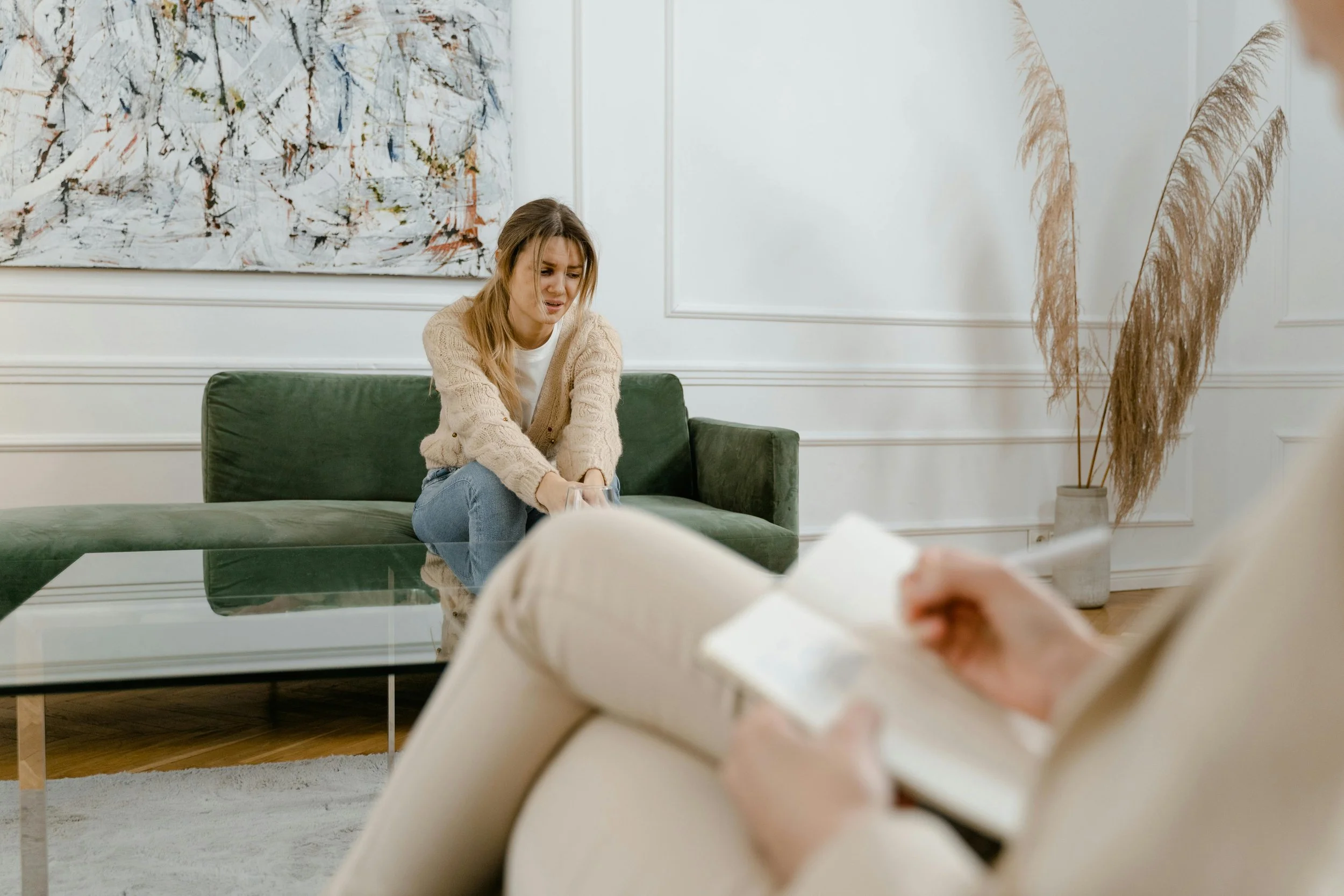 A young woman is sitting on a green sofa, appearing upset or distressed, in a therapy session with a therapist who is taking notes and reading from a notebook in a cozy, modern room with white walls, abstract art, and tall pampas grass in a vase.