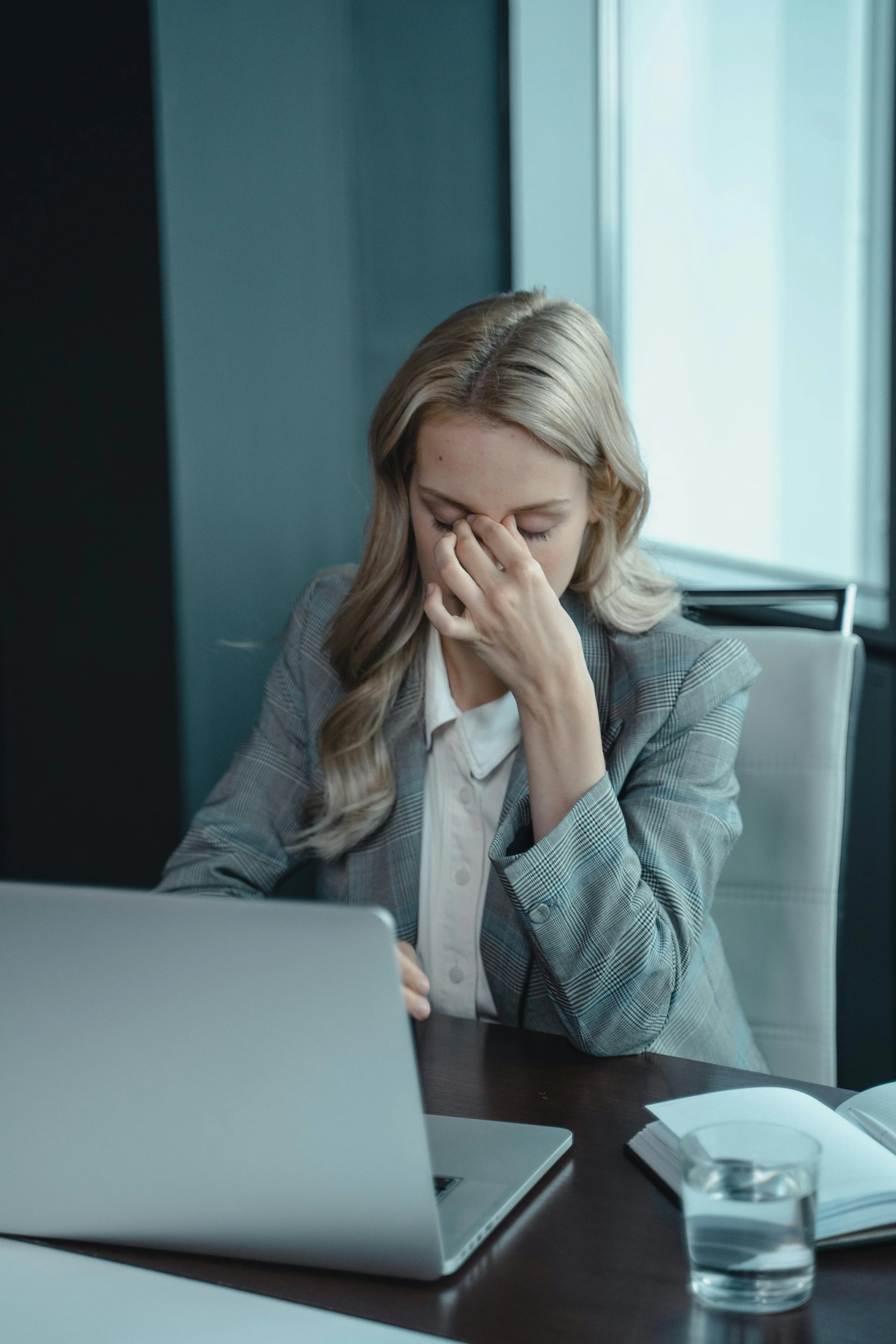 A woman sitting at a desk, appearing stressed or tired, with her hand covering her face, in front of a laptop, with a notebook and glass of water nearby.
