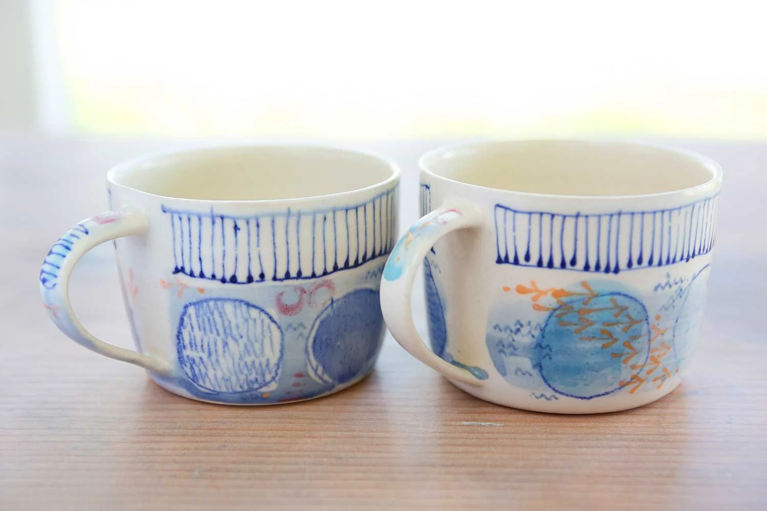 Two small batch ceramic mugs with blue and orange decorative patterns on a wooden surface.
