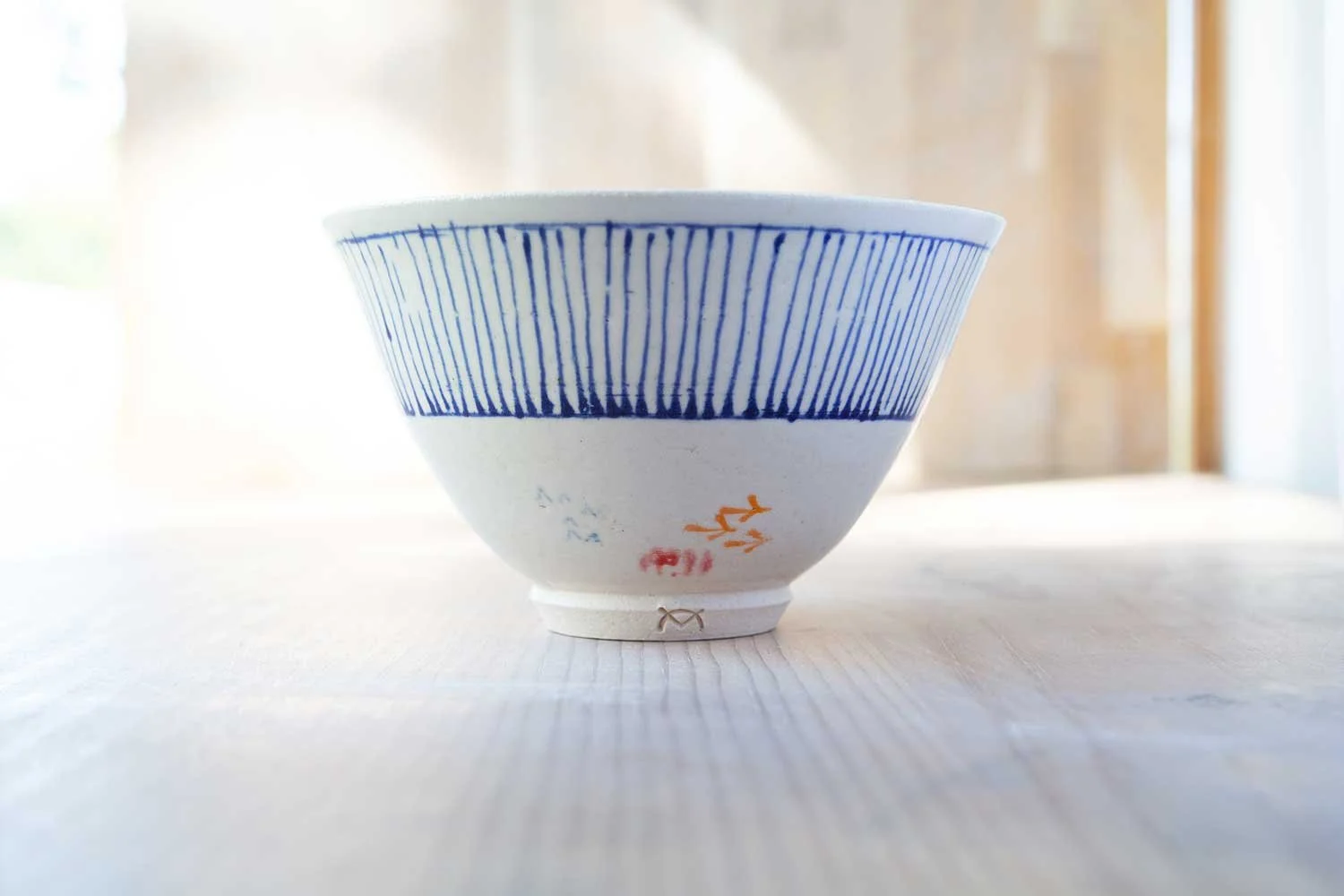 A white ceramic bowl with hand-painted blue vertical line patterns on the rim and some small, colorful designs at the base, placed on a wooden surface with a blurred background.