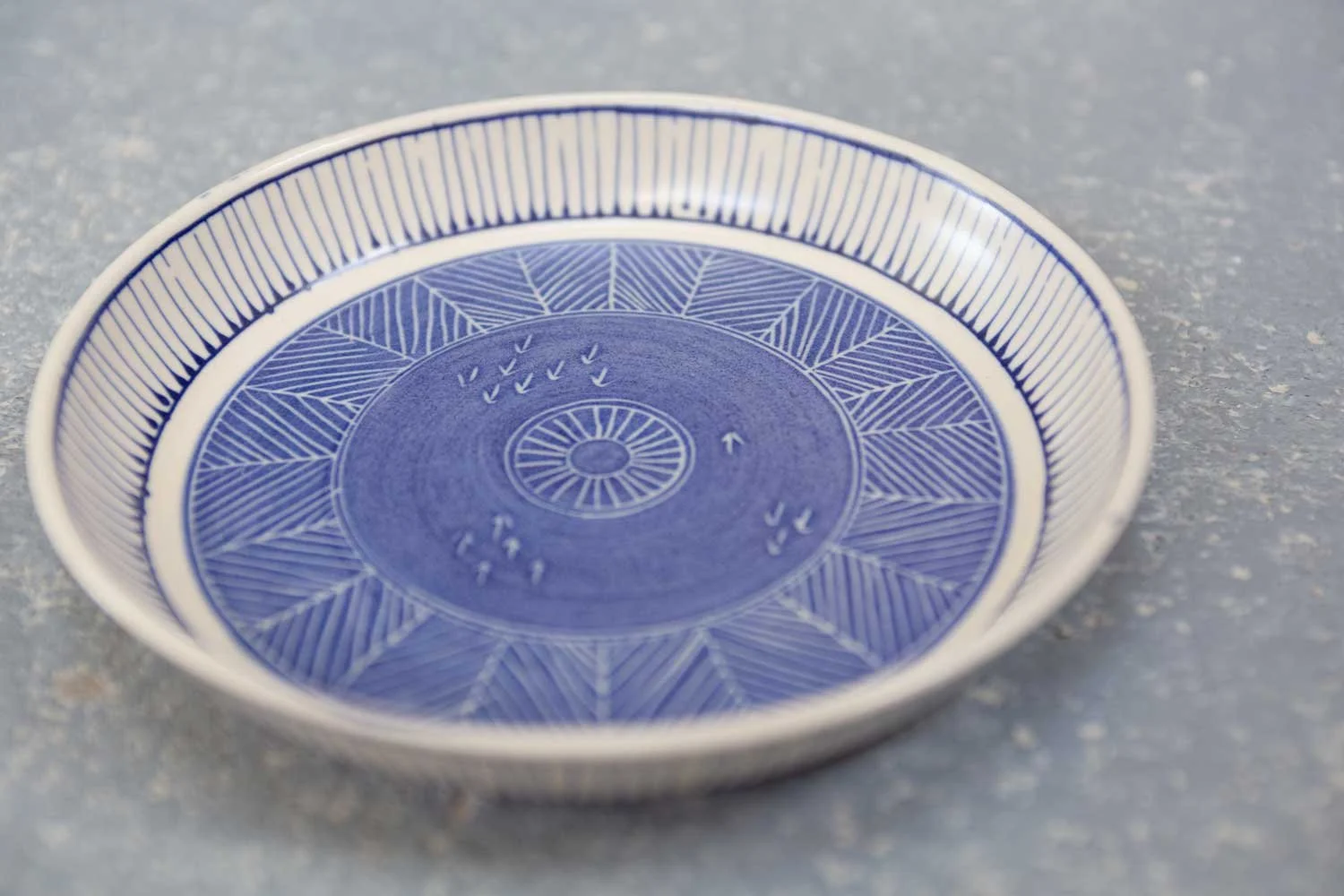 A Cornish blue and white patterned plate, decorated using the sgraffito technique, on a grey background.