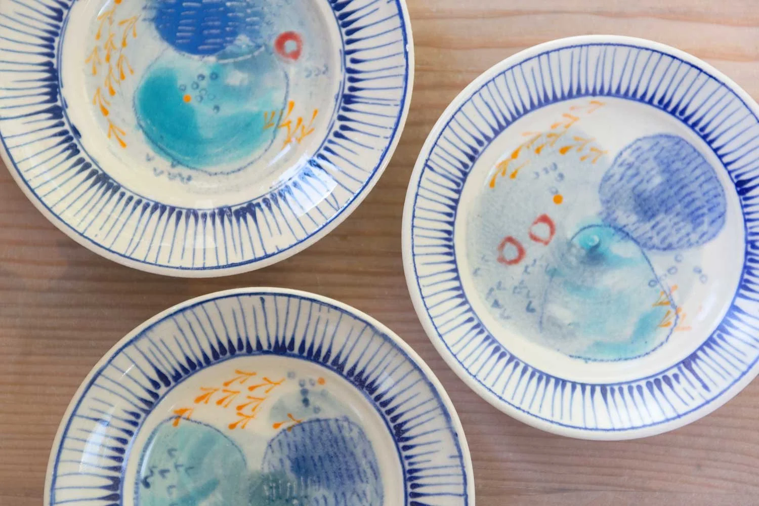 Three ceramic plates with blue, orange and red abstract patterns, viewed from above on a wooden surface.
