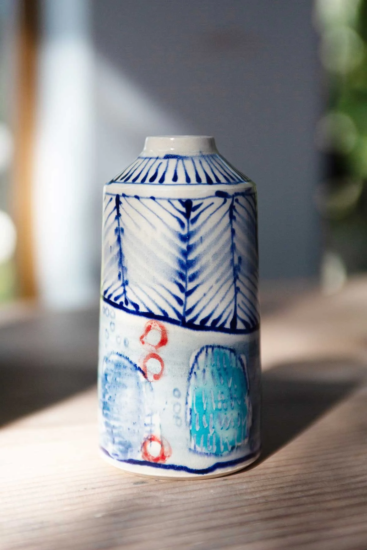 Decorative ceramic bud vase with blue, red, and turquoise abstract patterns on a wooden surface, lit up with natural light with a blurred background.