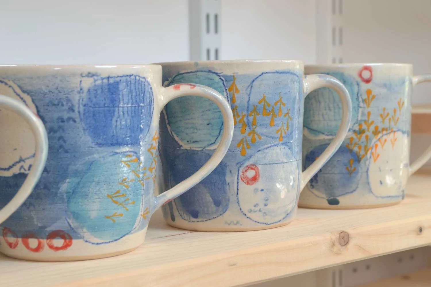 A row of unique ceramic mugs with abstract blue, yellow, and red patterns, created by artisan Claire Lean.