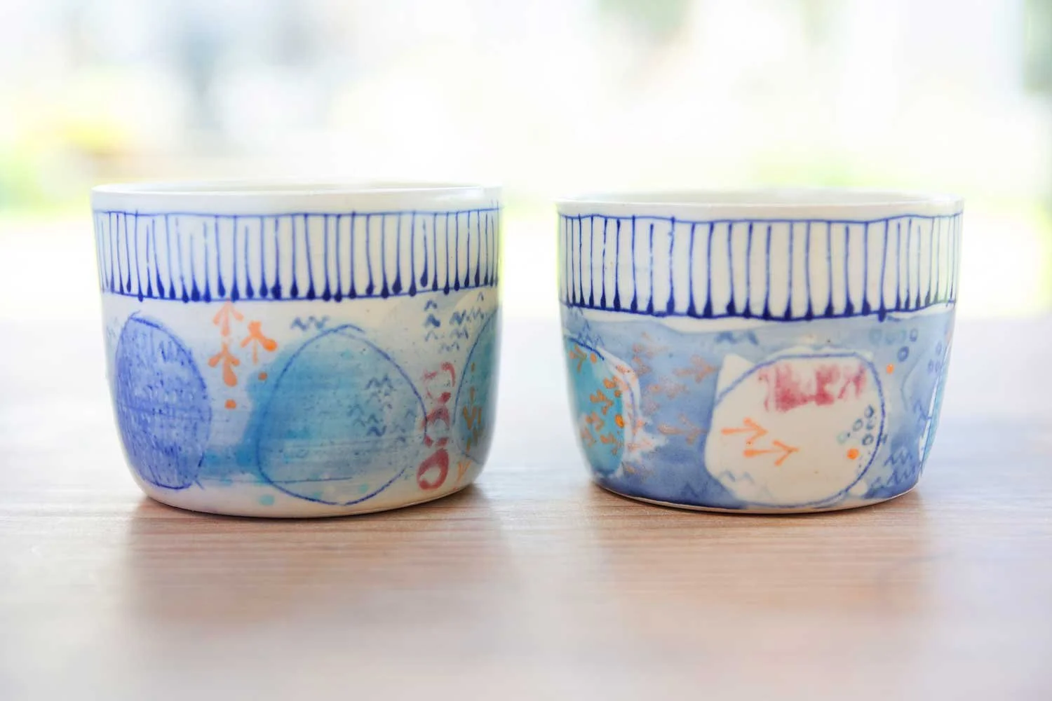 Two colourful hand decorated ceramic bowls placed side by side on a wooden surface with a blurred natural background.