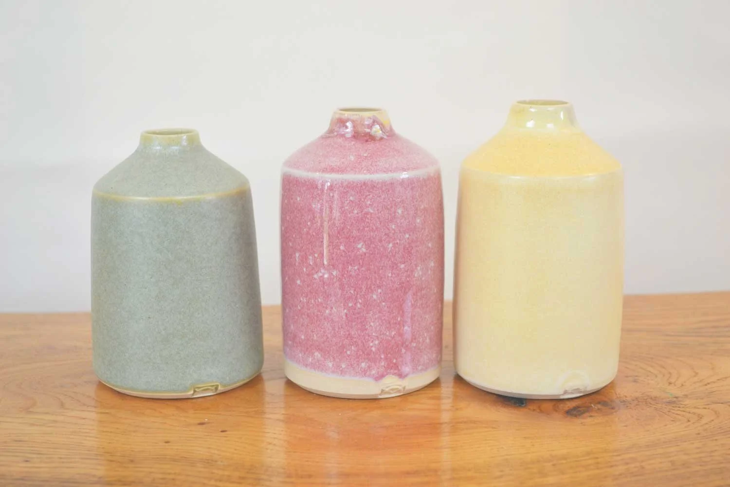 Three ceramic vases in coordinated tonal shades of light green, pink, and yellow, arranged on a wooden surface against a white background.