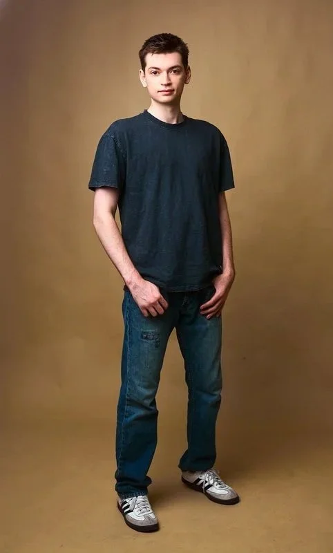 Young man with short dark hair wearing a dark t-shirt, jeans, and sneakers standing against a plain background.