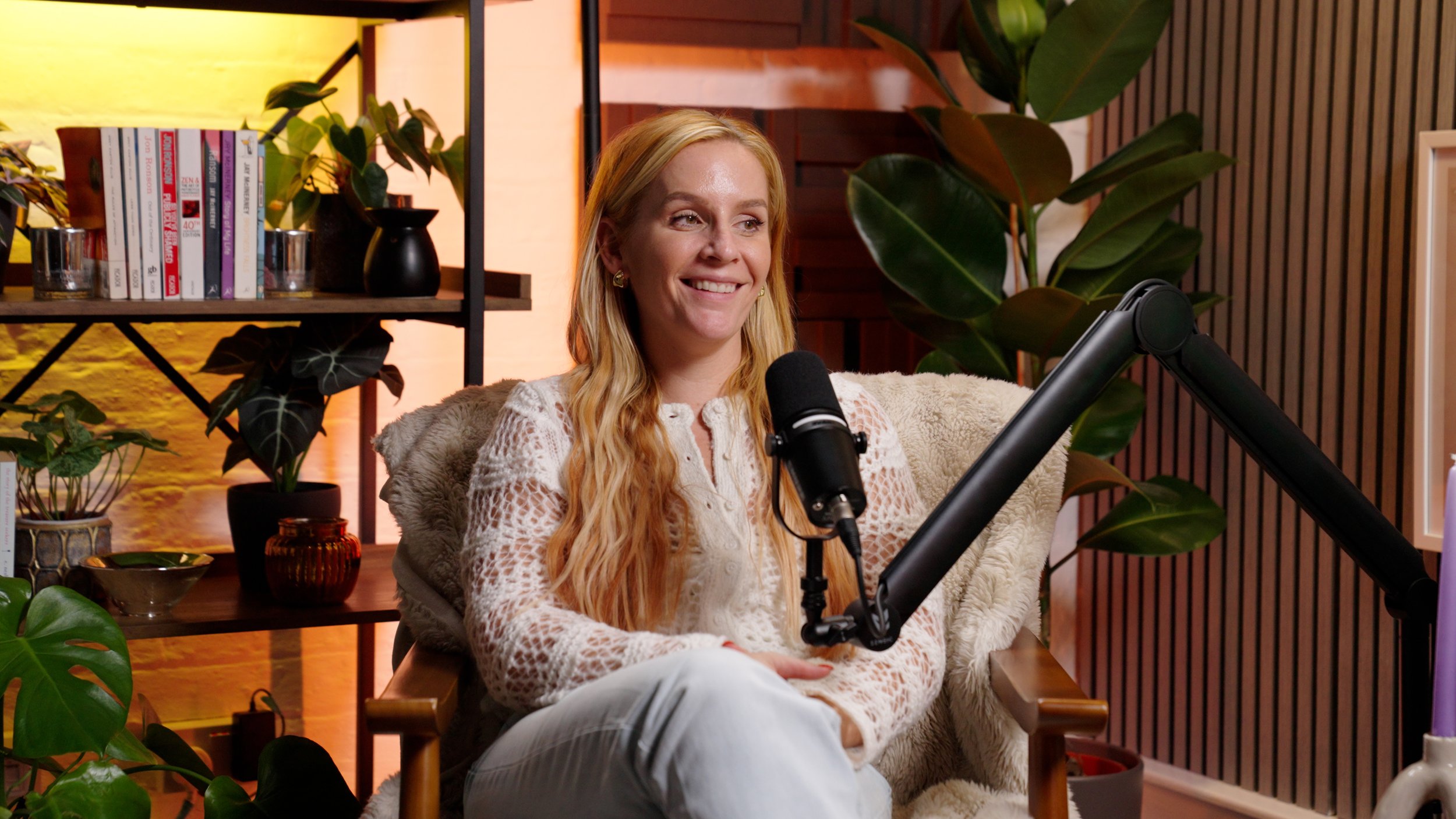 A woman sitting in front of a microphone, smiling, surrounded by plants and shelves with books and decorative items.