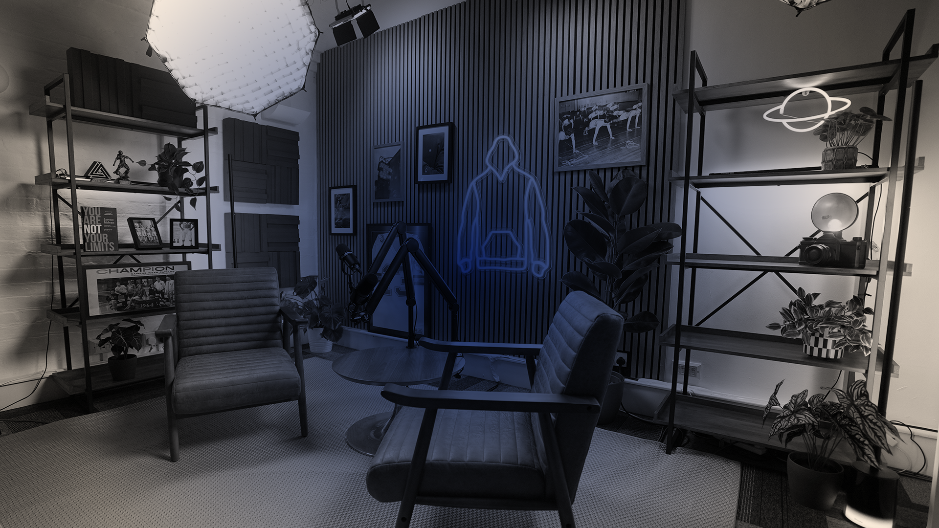 A modern office or studio room with two shelves filled with plants, photos, and decor, a neon outline of a person with a book on the wall, and a camera on a shelf. There are two chairs, a round table, and a large umbrella light, with framed pictures and artwork on the walls.