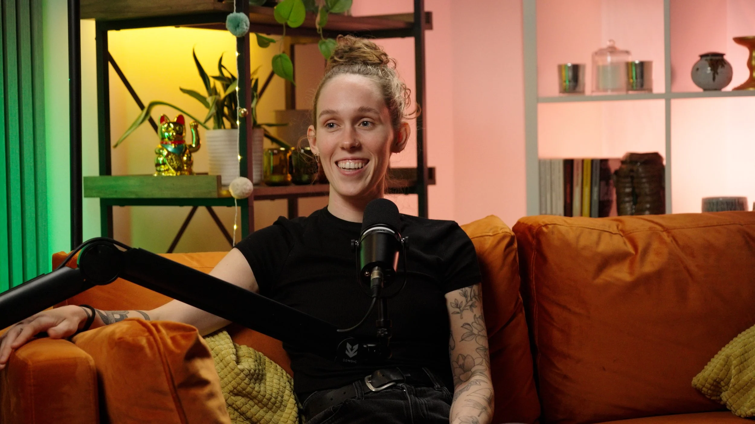A young woman with blonde hair tied up in a bun, wearing a black shirt, is sitting on an orange couch with yellow cushions, smiling during a podcast recording with a microphone in front of her. The background features a bookshelf with plants, books, and decorative items.