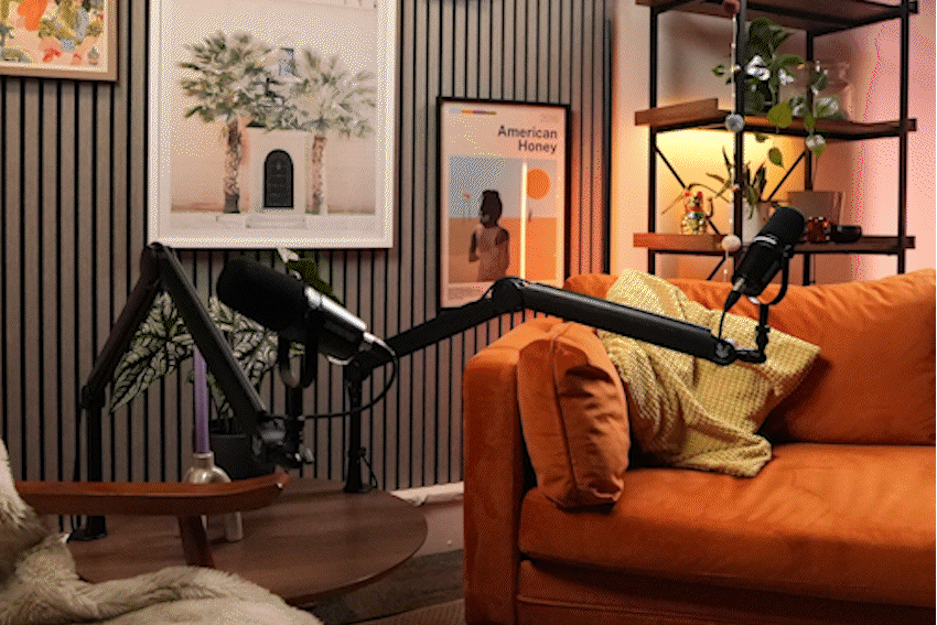 A cozy living room setup with a burnt orange sofa, pillows, and a black microphone arm extending over the sofa, with a recording microphone attached. The background features framed artwork, a black textured wall, a wooden shelf with plants and decor, and a side table with a candle.
