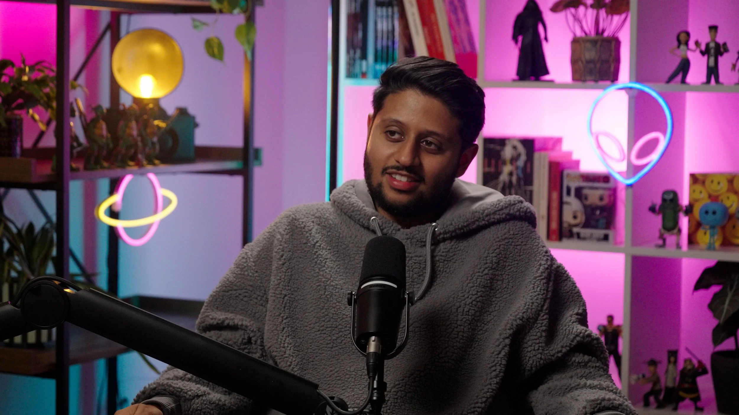 A young man with a beard and short dark hair, wearing a gray hoodie, sitting in front of a microphone in a colorful room with pink and blue LED lights and shelves filled with books, toys, and decorations including neon alien heads and planets.