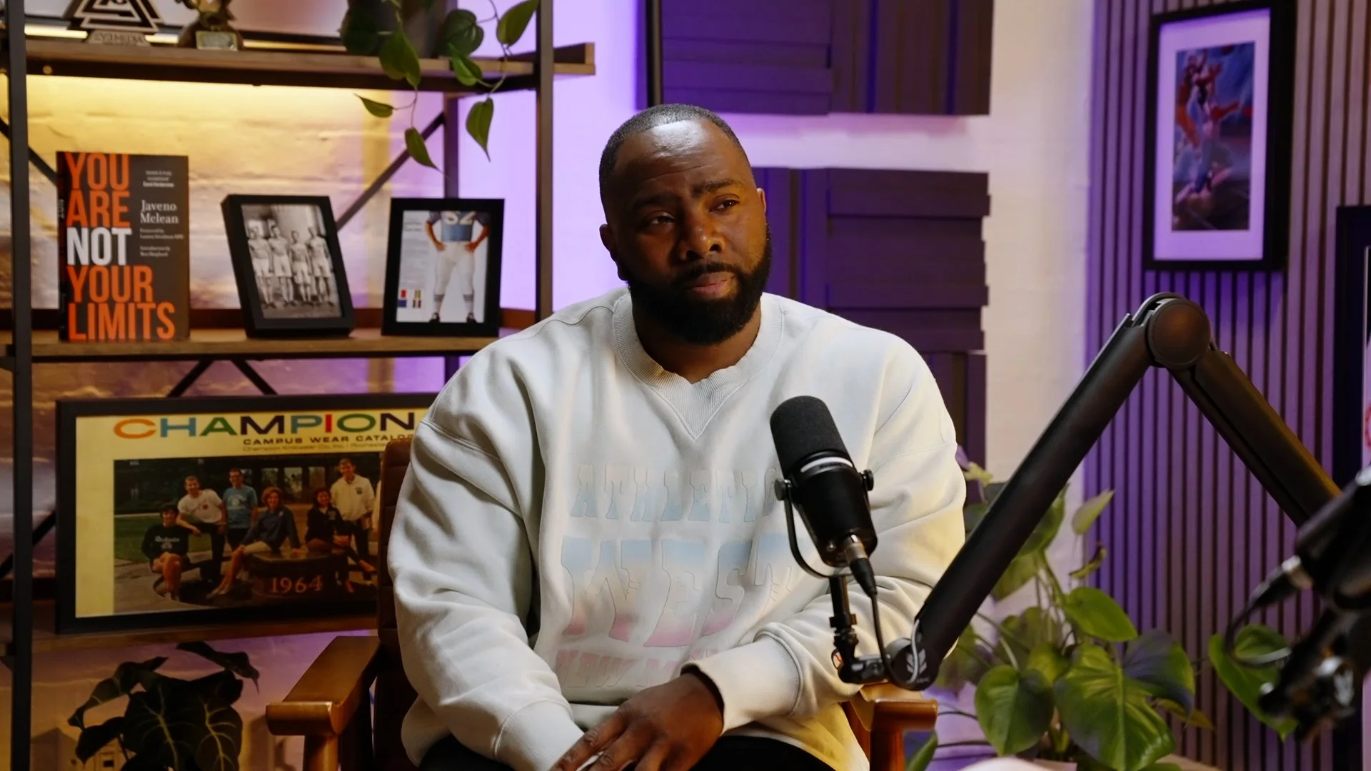A man sitting in a chair in a recording studio with a microphone in front of him. The background features shelves with framed photos, books, and a poster, along with plants and decorative panels.