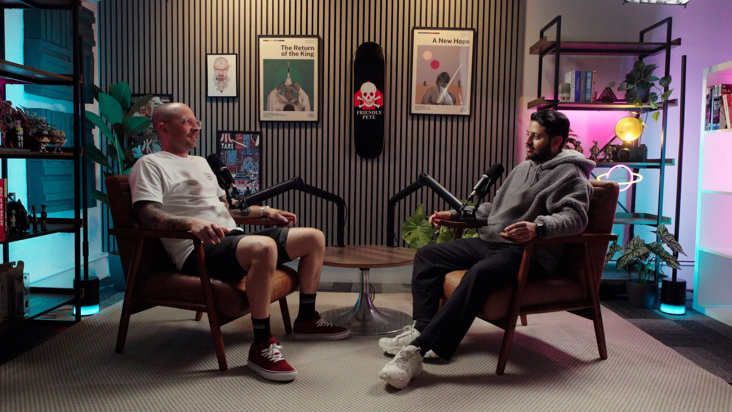 Two men sit in chairs facing each other in a recording studio with microphones, decorated with posters, plants, and colored lights.