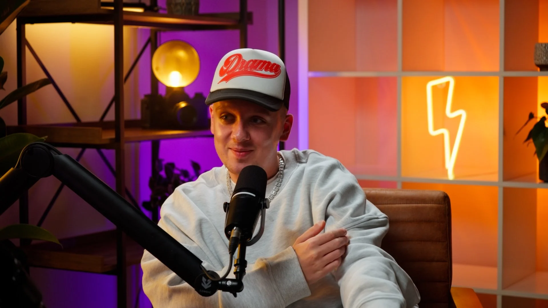 A young man wearing a white sweatshirt, a silver chain, and a white cap with red lettering sitting at a desk with a microphone, in a colorful studio with neon lighting and decor.