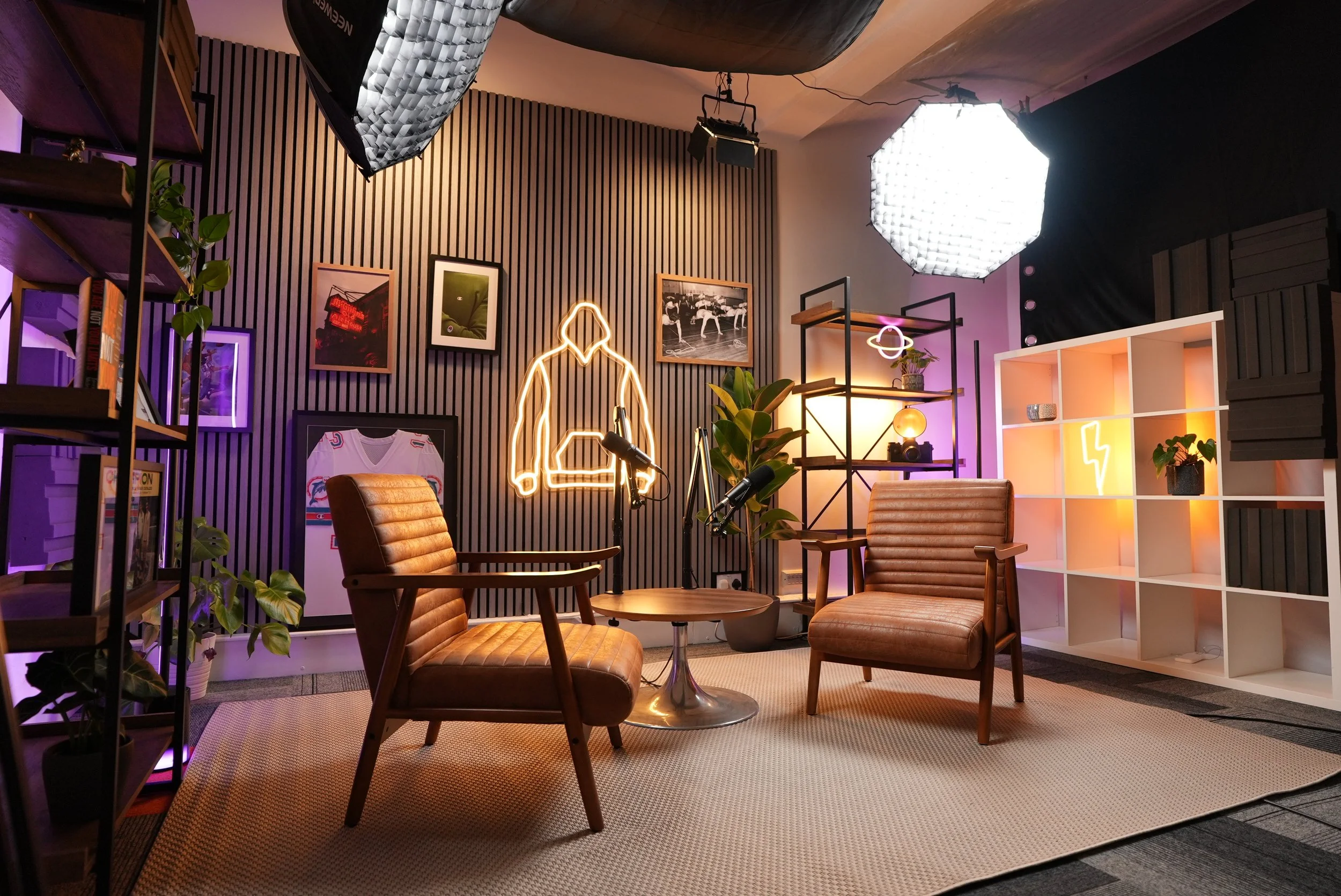A modern studio with two vintage-style leather armchairs around a small round wooden table. The background features a black slatted wall with framed pictures and neon light art shapes, including a glowing insignia of a shirt and a human outline. To the right, there is a white shelving unit with plants, decorative items, and a neon lightning bolt. Lighting includes a large octagonal softbox, a smaller pink neon circle, and a globe light, creating a cozy, creative atmosphere.