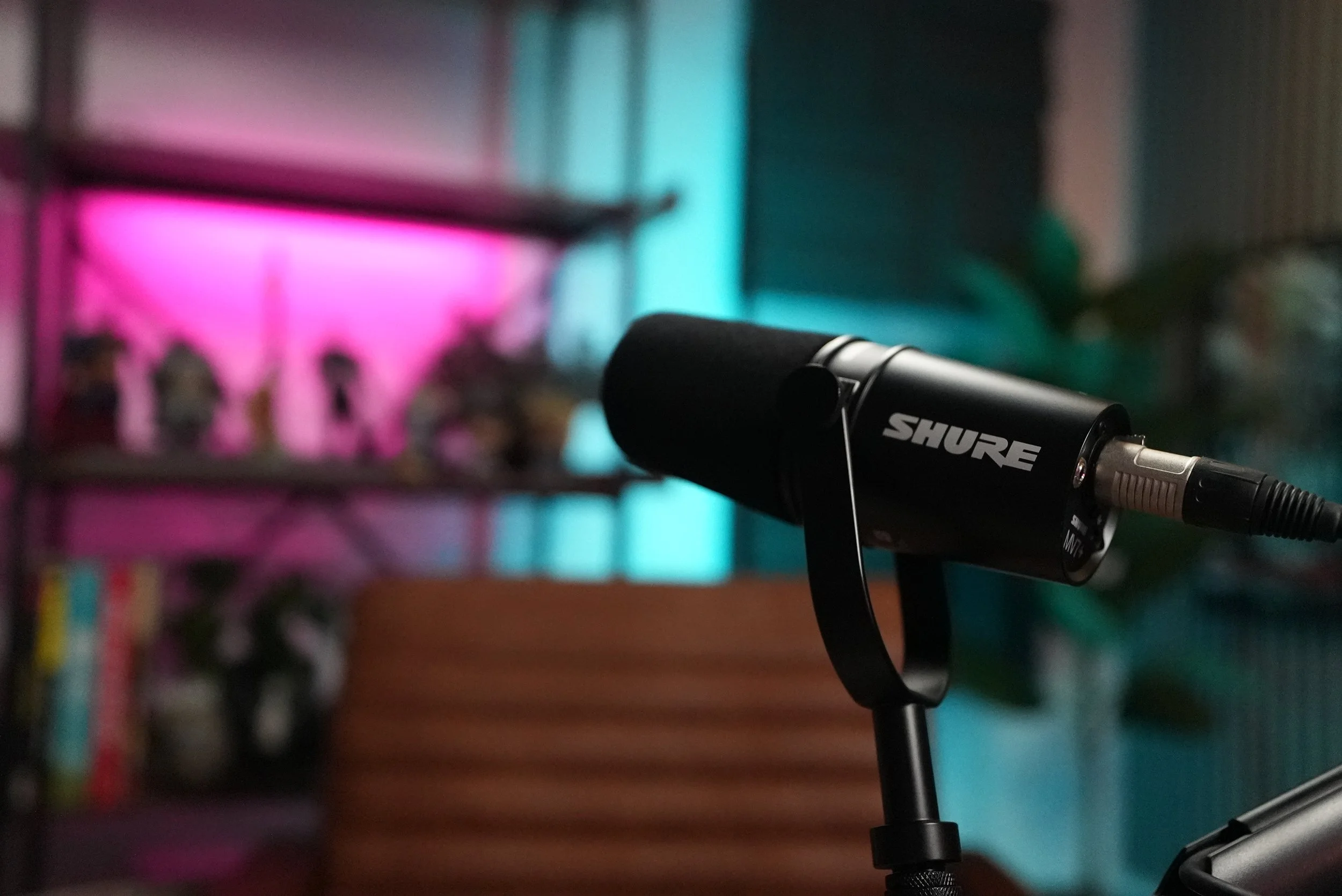 Close-up of a black Shure microphone mounted on a stand in a room with colorful blurred background including pink and blue lighting.