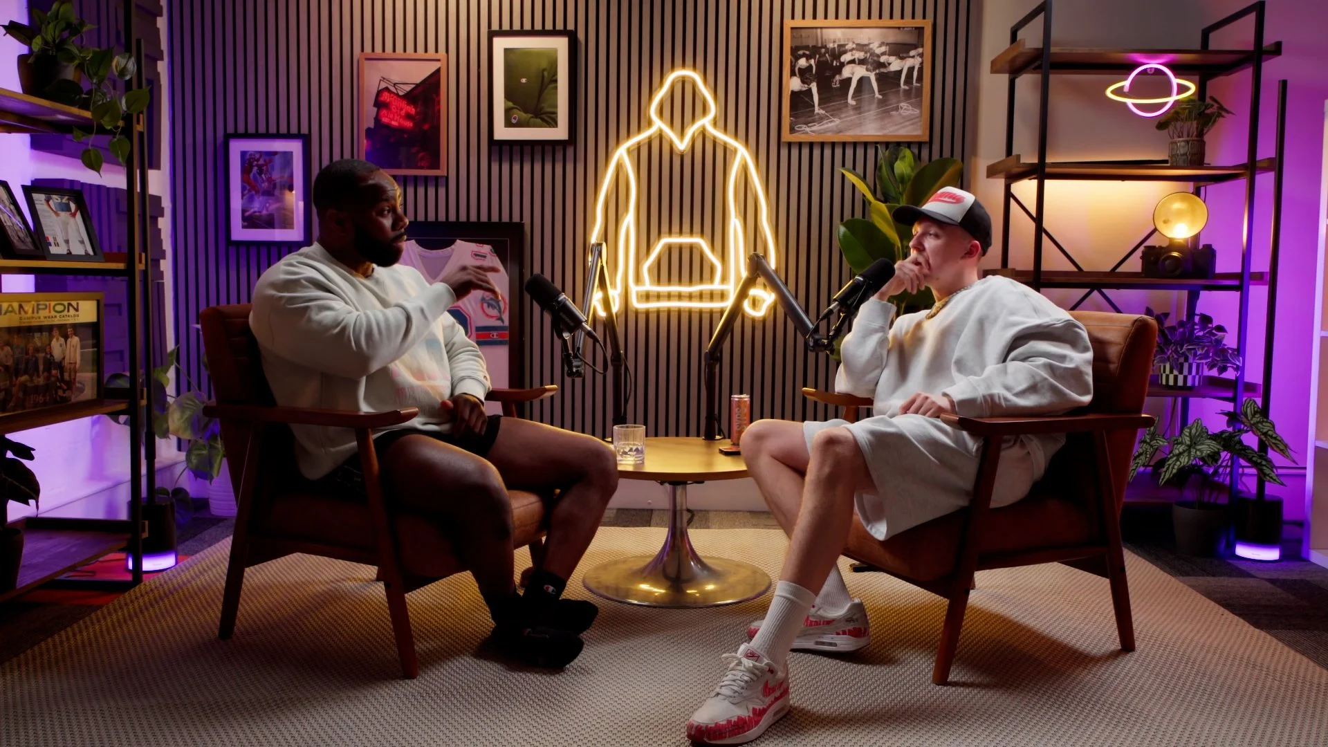 Two men sit across from each other in a podcast room with microphones, wearing casual white sweatshirts. Behind them is a background with black and purple lighting, framed photos, and a neon outline of a person holding a basketball. There are shelves with books and plants on either side.