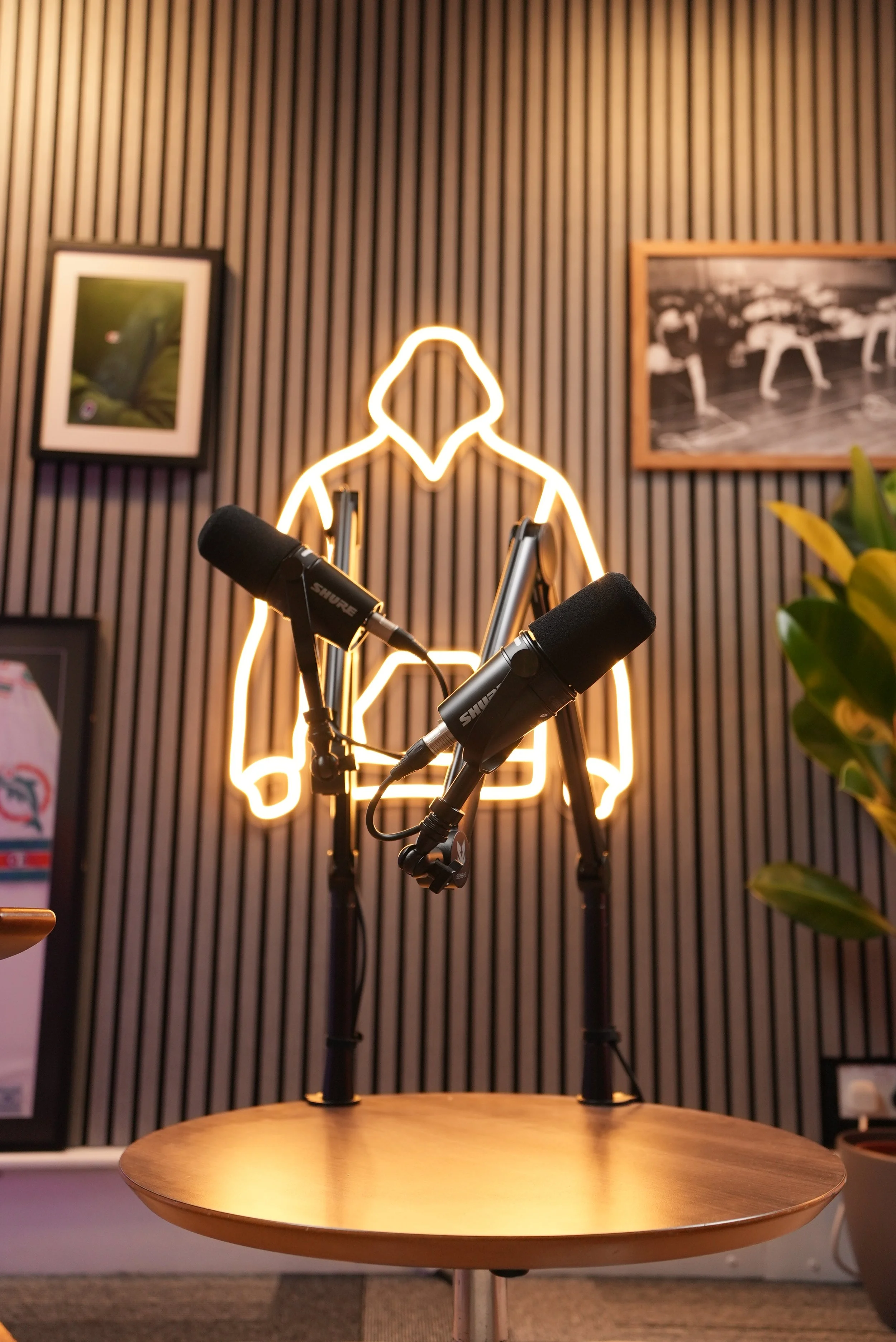Microphone setup in front of a neon outline of a person sitting on a chair, with framed photos on a ribbed wall behind.