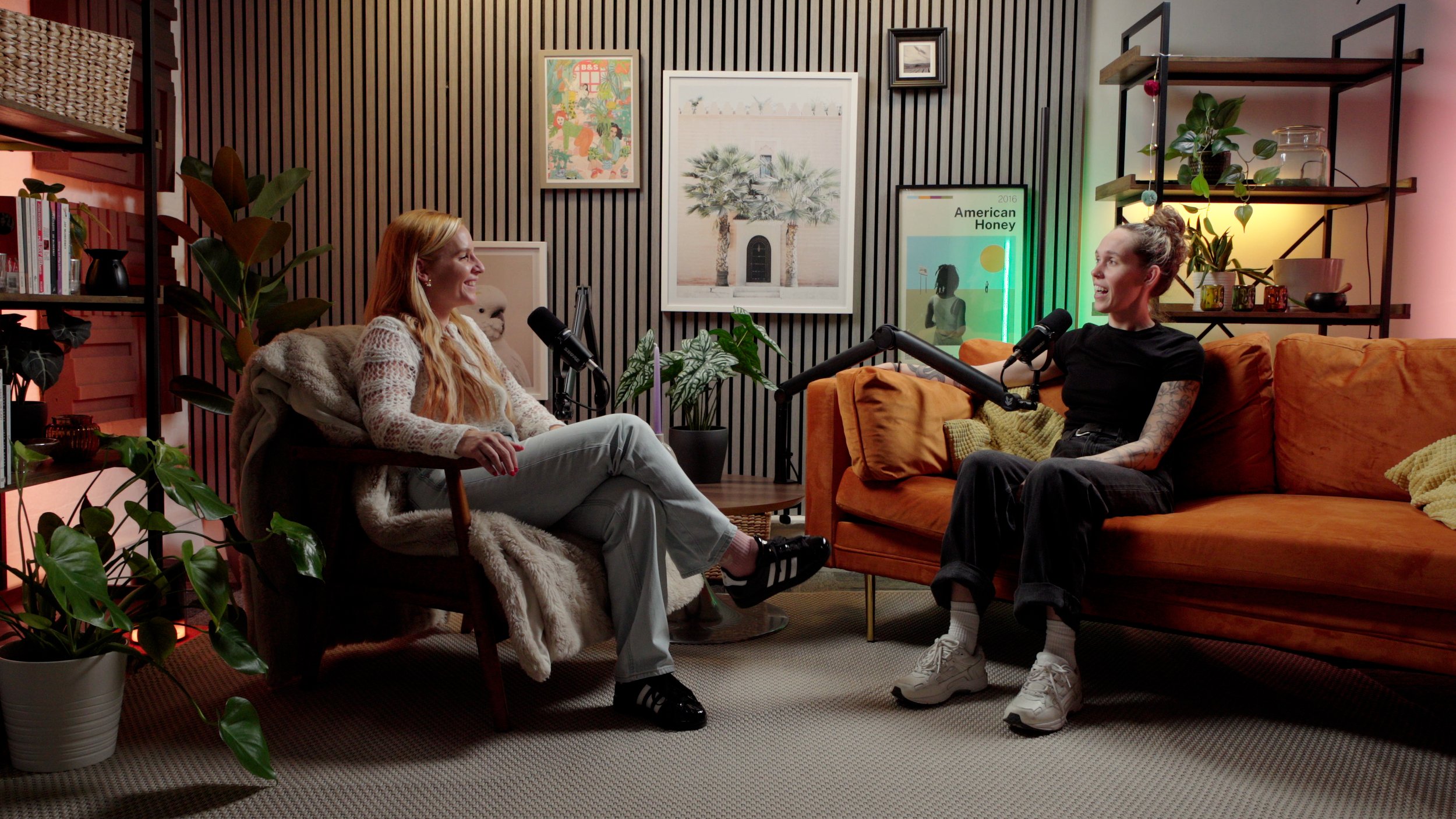 Two women having a conversation in a cozy, modern living room with black and orange furniture, plants, and wall art, each speaking into a microphone.