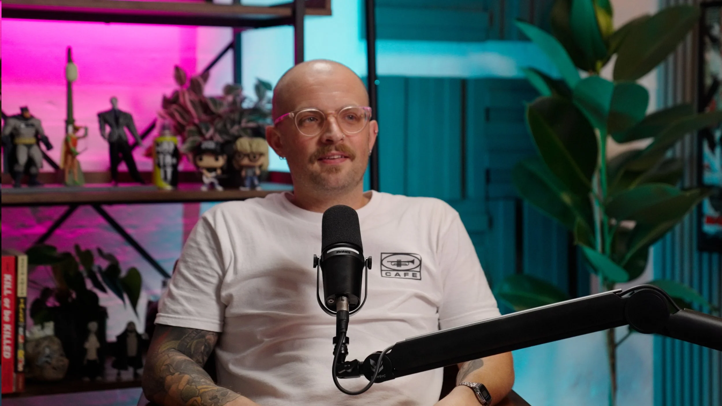 A man with glasses and a mustache, wearing a white T-shirt with a logo, speaking into a microphone during a podcast or interview. Behind him are shelves with action figures and books, with colorful lighting and indoor plants.