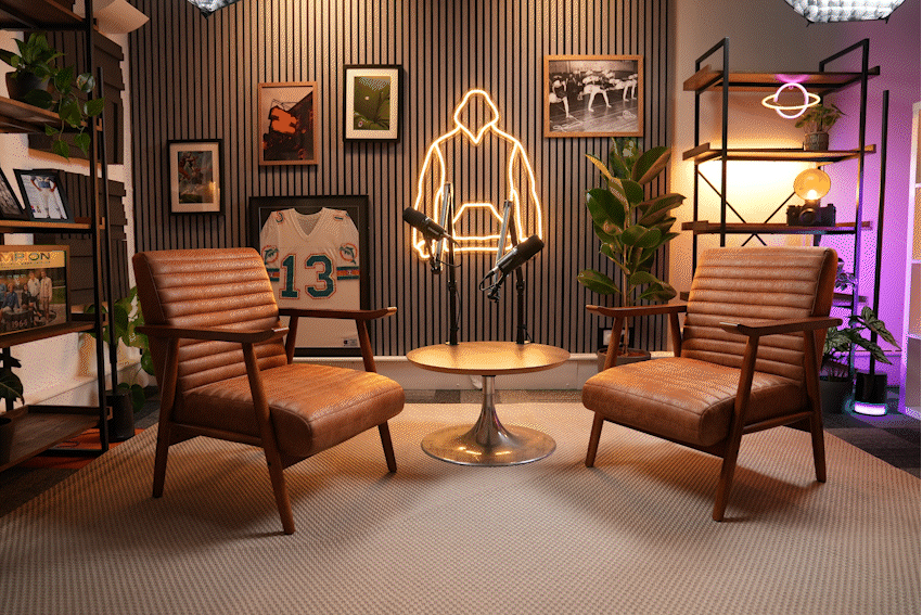 A modern podcast or interview setup with two brown leather chairs facing each other, a round table in between, and a wall decorated with framed photos and artwork. There is a neon outline of a person on the wall behind the table, and two microphones are positioned on the table. The room is decorated with plants and ambient lighting.