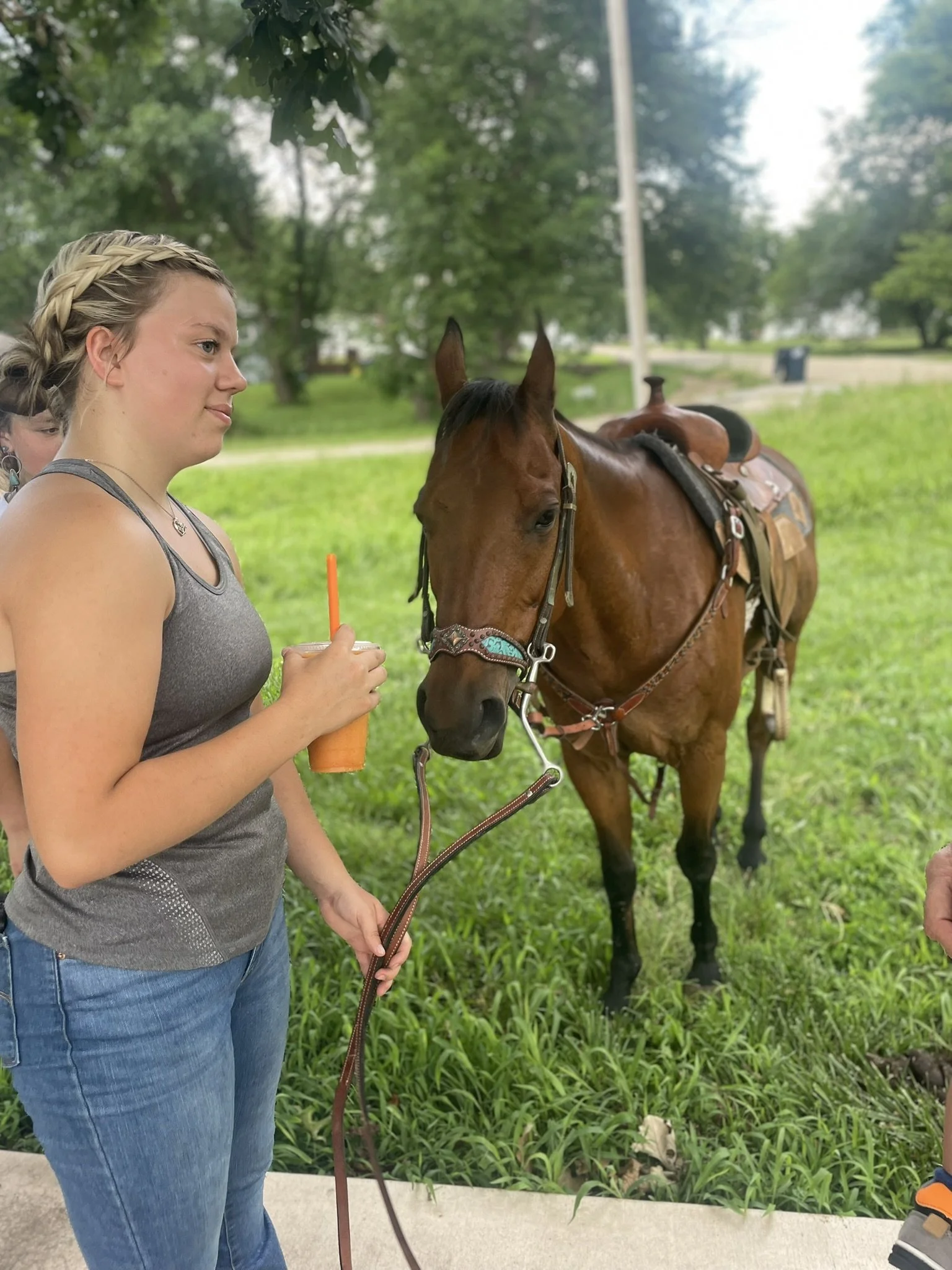 Elm Creek Beef A young woman with blonde hair in a braid holding a brown horse by a lead rope, with her left hand, while holding an orange beverage in her right hand, outdoors on a grassy area with trees in the background.