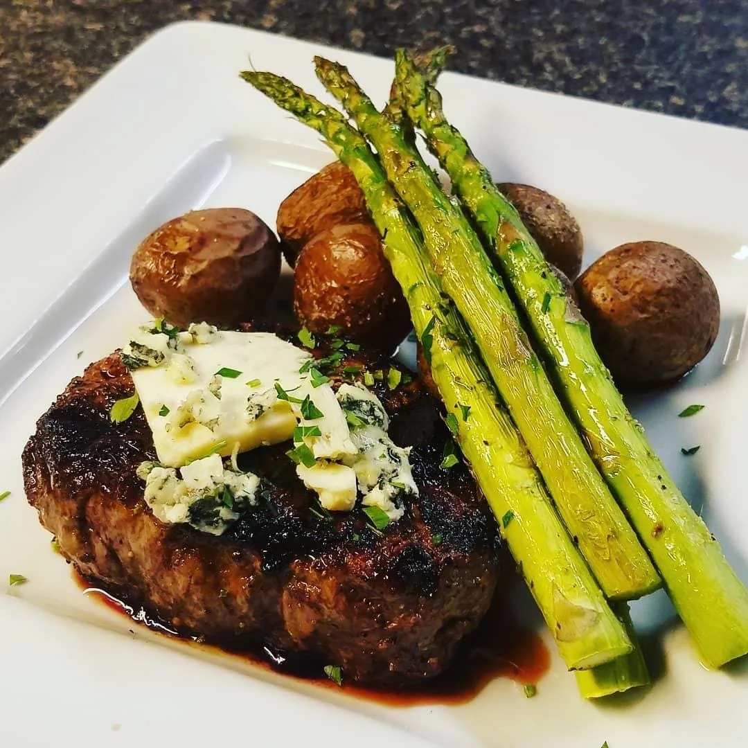 Elm Creek Beef Steak topped with blue cheese, accompanied by roasted potatoes, grilled asparagus, and mushrooms on a white plate.