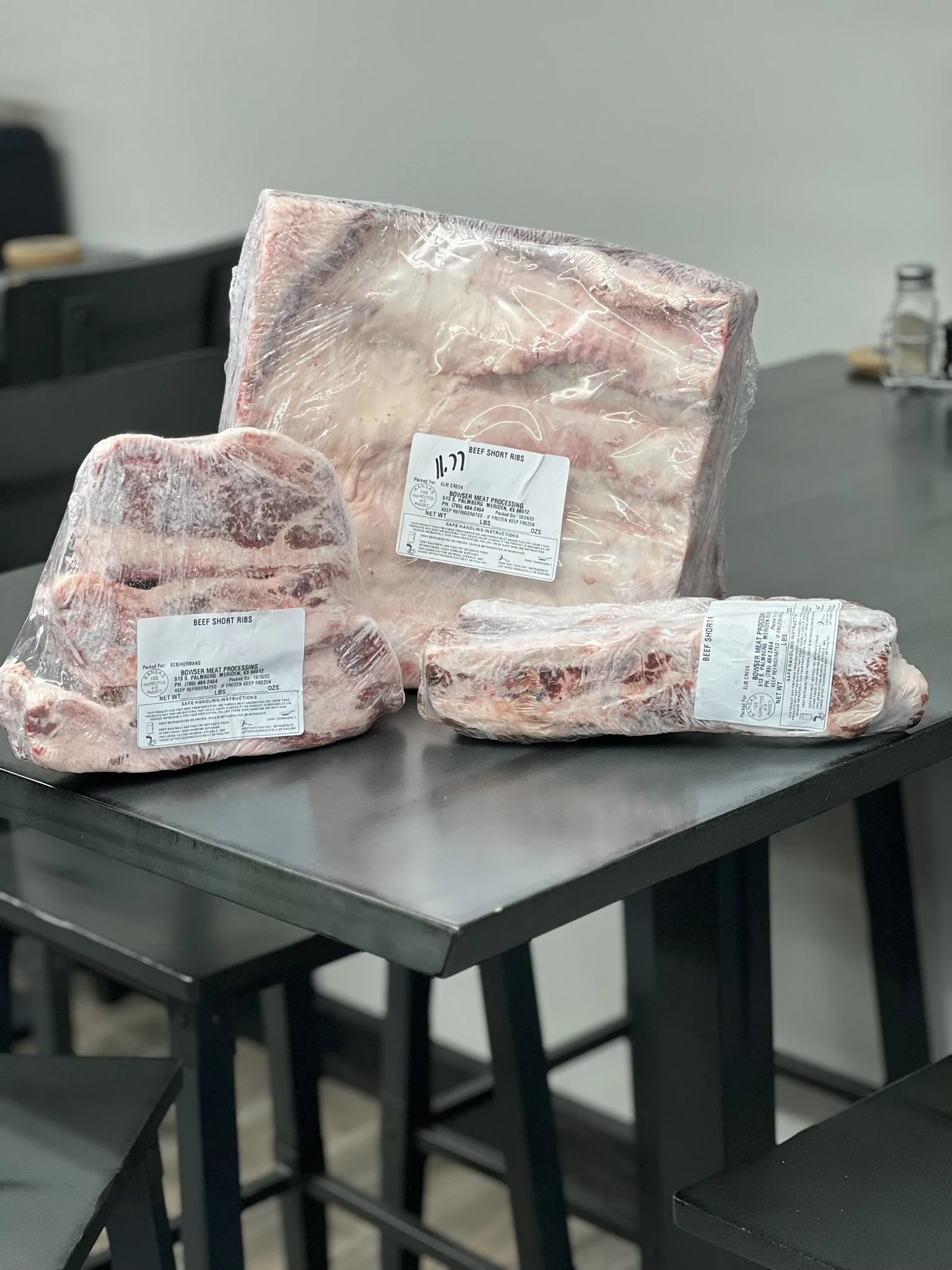 Elm Creek Beef Three packages of beef short ribs on a black table, with one large piece wrapped in plastic, a smaller piece, and a smaller package. All packages have white labels.