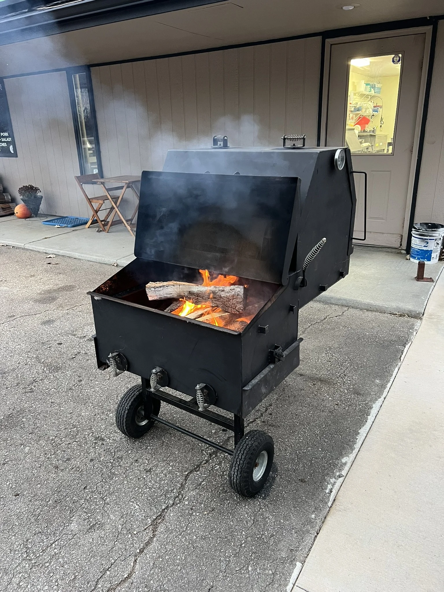 Elm Creek Beef A black outdoor smoker with a lid open, revealing burning logs and smoke, situated on a paved surface outside a building with beige walls and a windowed door.