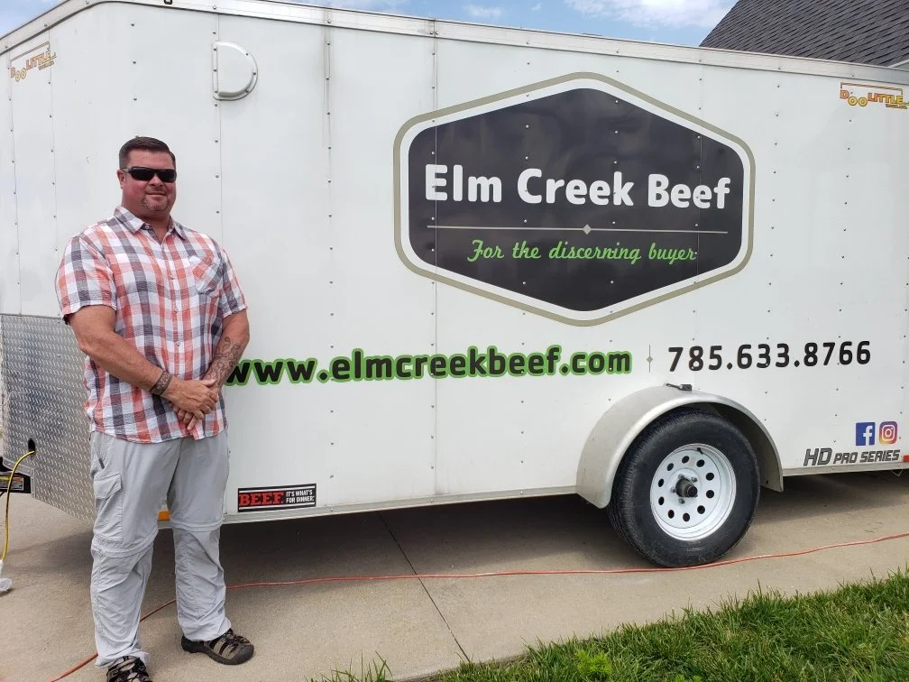 Elm Creek Beef Man in sunglasses standing next to a mobile trailer for Elm Creek Beef, with website and contact information on the trailer.