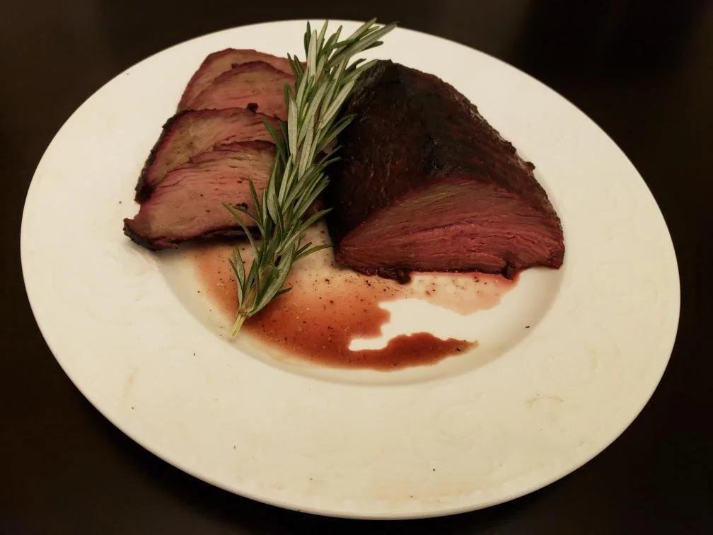 Elm Creek Beef Sliced cooked steak with a sprig of rosemary and some sauce on a white plate.