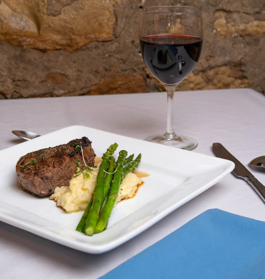Elm Creek Beef A plate with a cooked steak, mashed potatoes, and asparagus with a glass of red wine on a table against a brick wall.