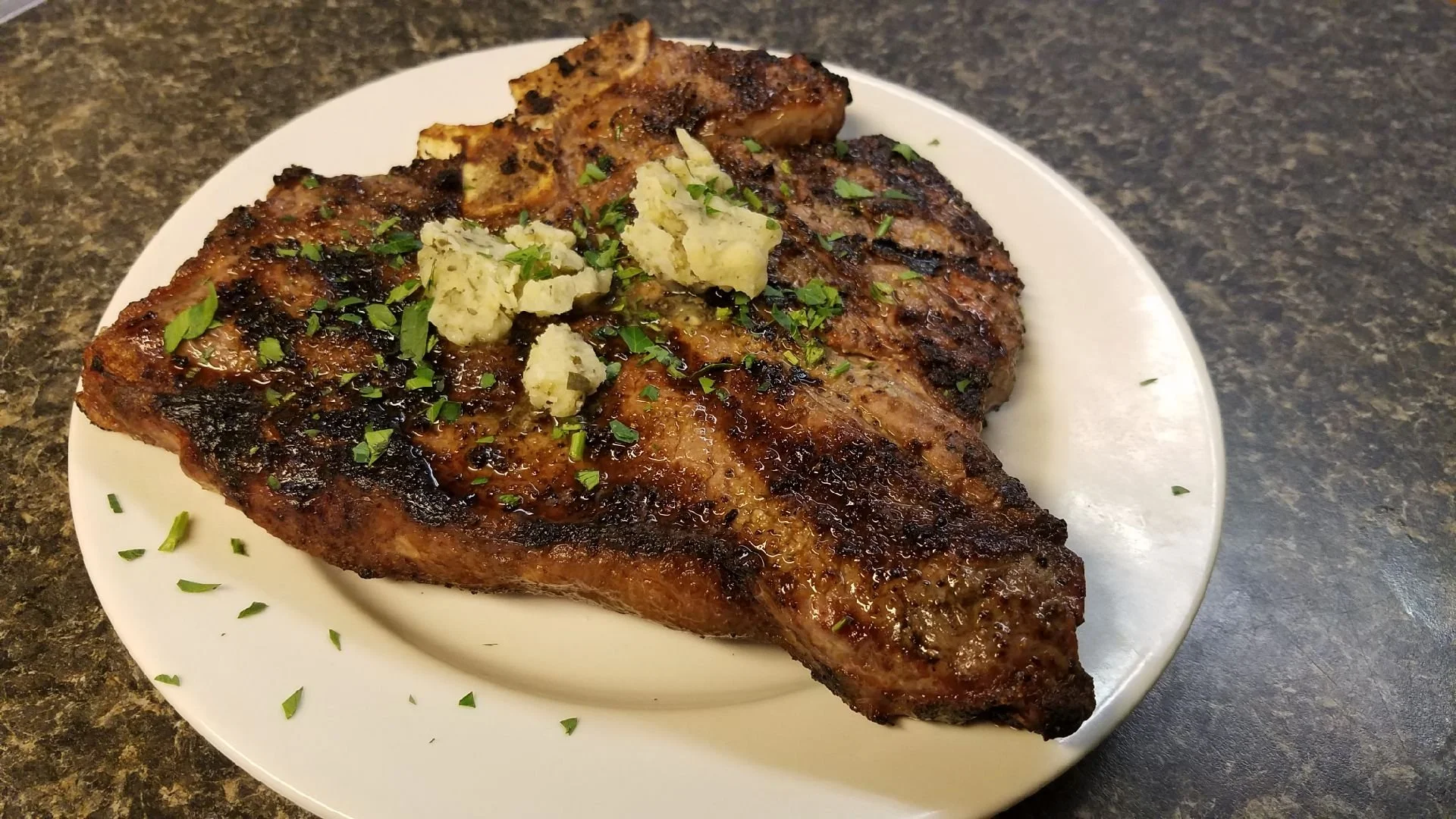 Elm Creek Beef Grilled steak with blue cheese crumbles and chopped herbs on a white plate over a dark countertop.