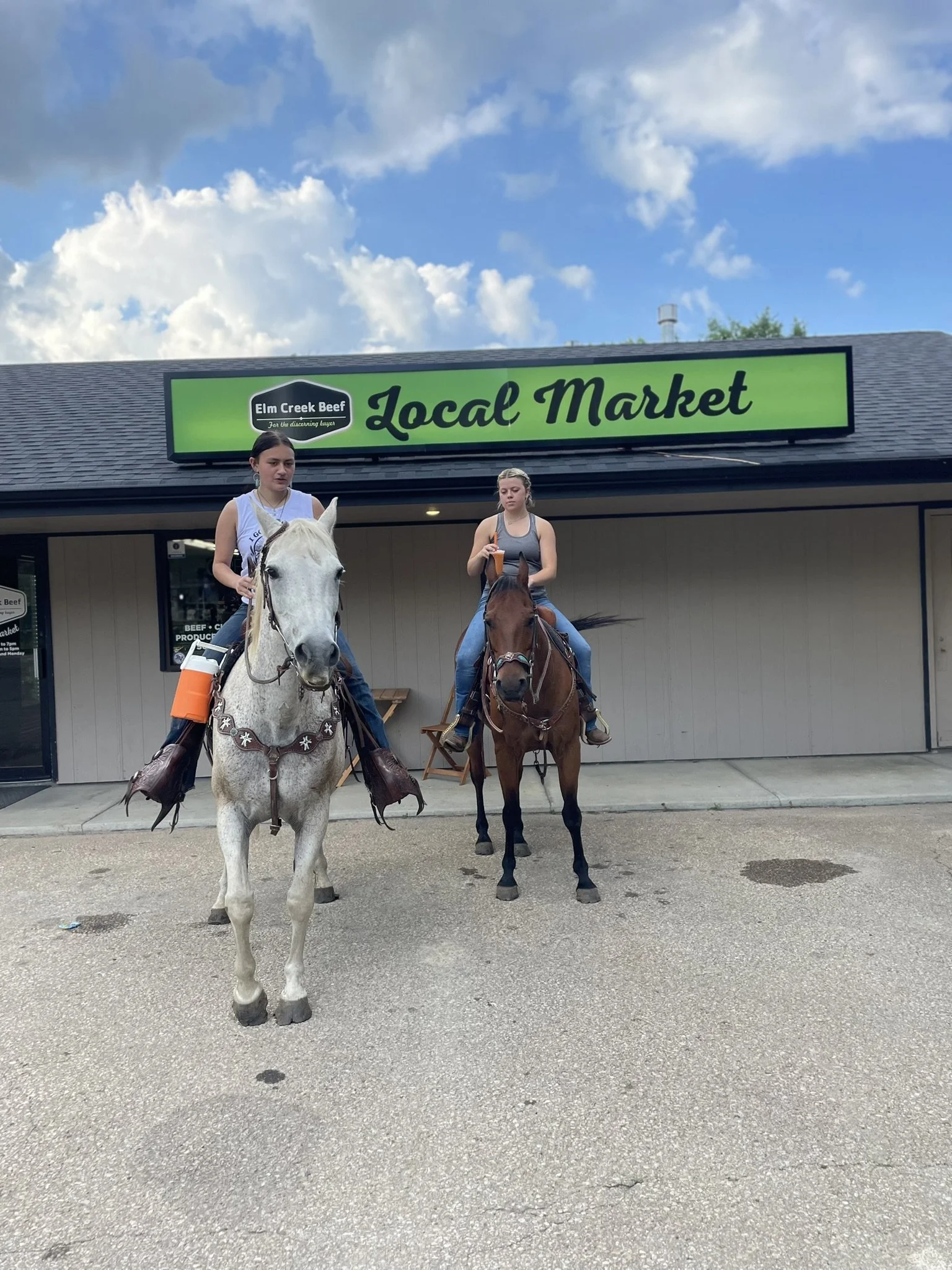 Elm Creek Beef Two young women riding horses in front of Elm Creek Beef Local Market, with a partly cloudy sky overhead.