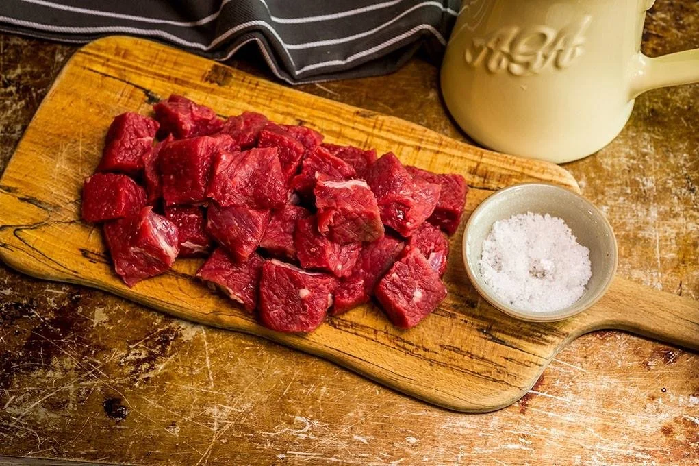 Elm Creek Beef Chopped raw beef on a wooden cutting board alongside a small bowl of coarse salt, with a beige mug in the background on a rustic wooden surface.