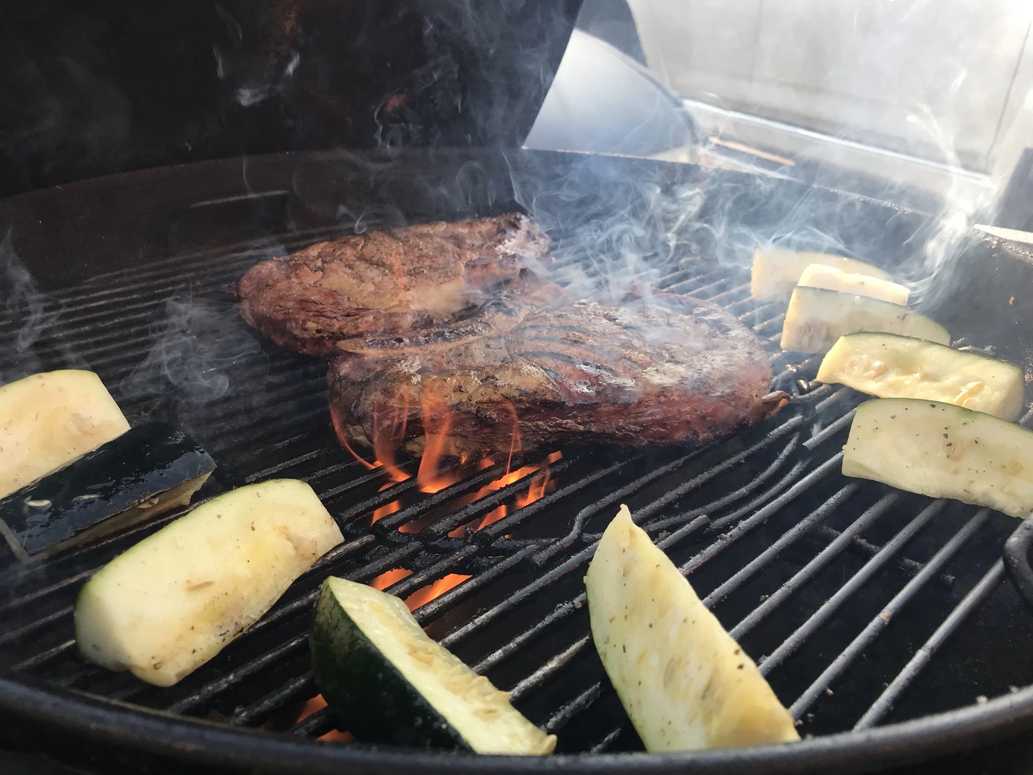 Elm Creek Beef Meat patties grilling on a barbecue with zucchini slices, some with char marks, and smoke rising from the grill.
